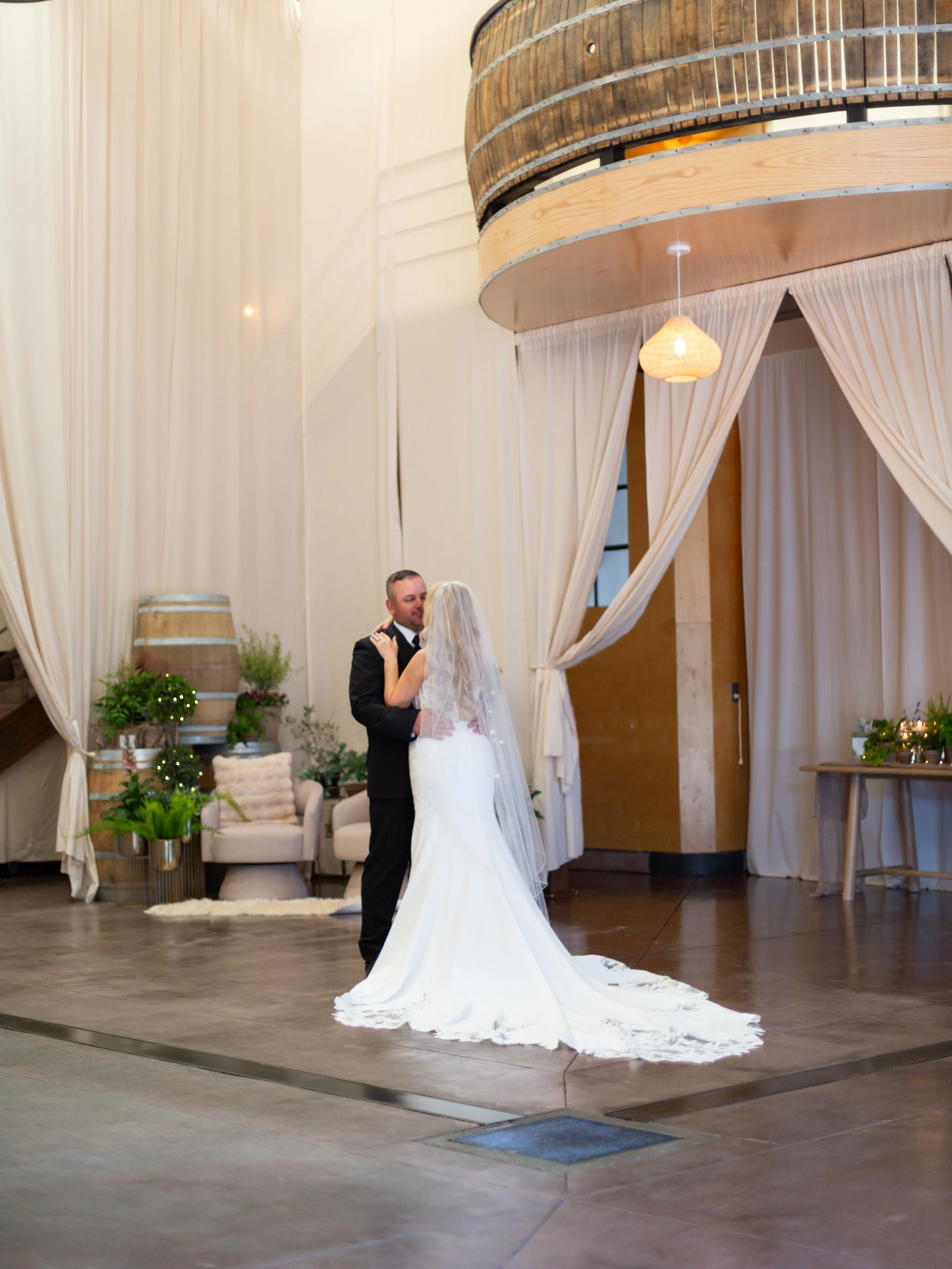 A bride and groom dancing during their wedding reception, with the bride wearing a long white wedding gown with a train and the groom in a black tuxedo, in a decorated indoor venue with beige curtains, plants, and cozy seating.