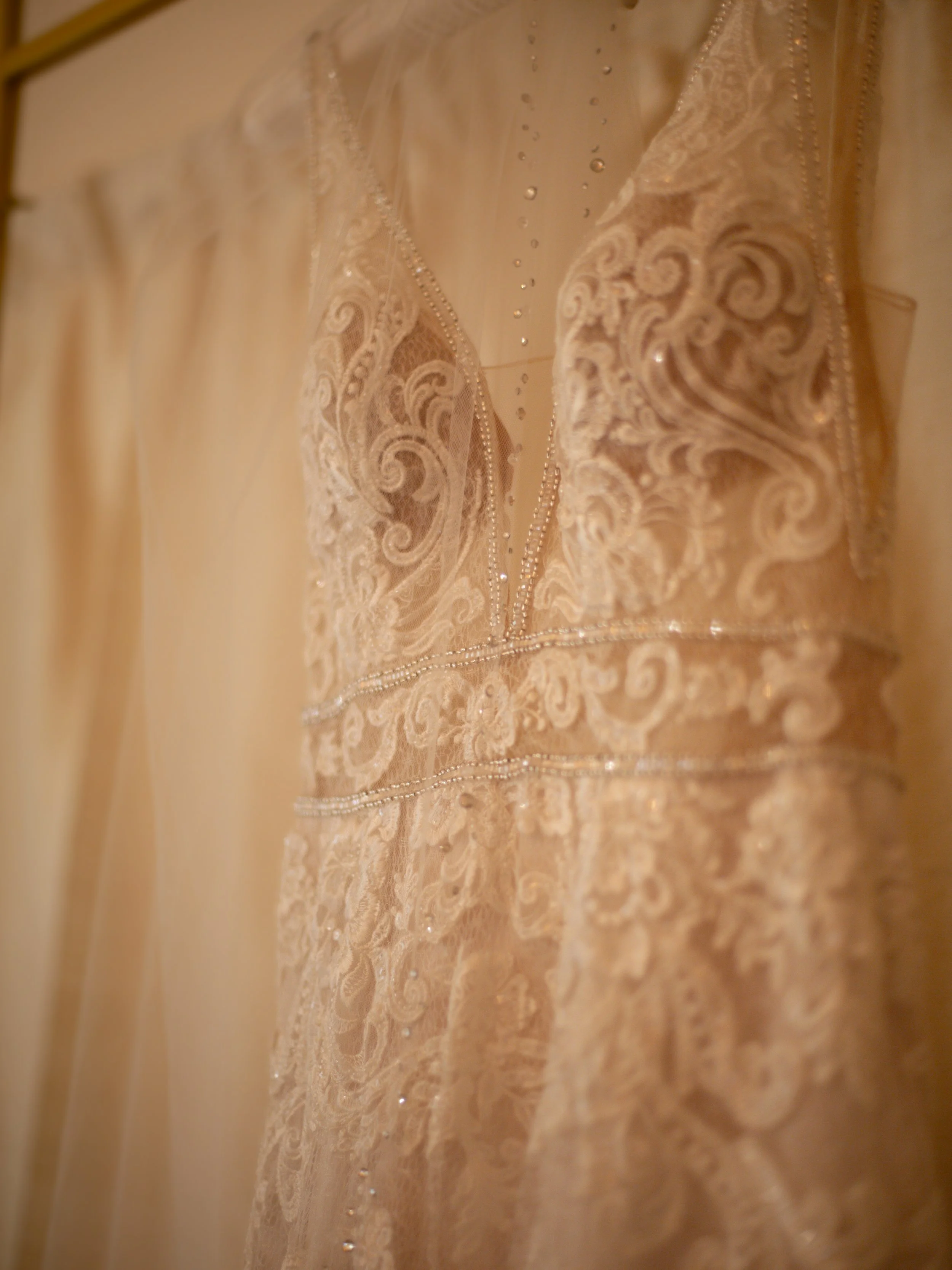 Close-up of a beige lace fabric with intricate swirling patterns and small beads, likely part of a wedding dress.