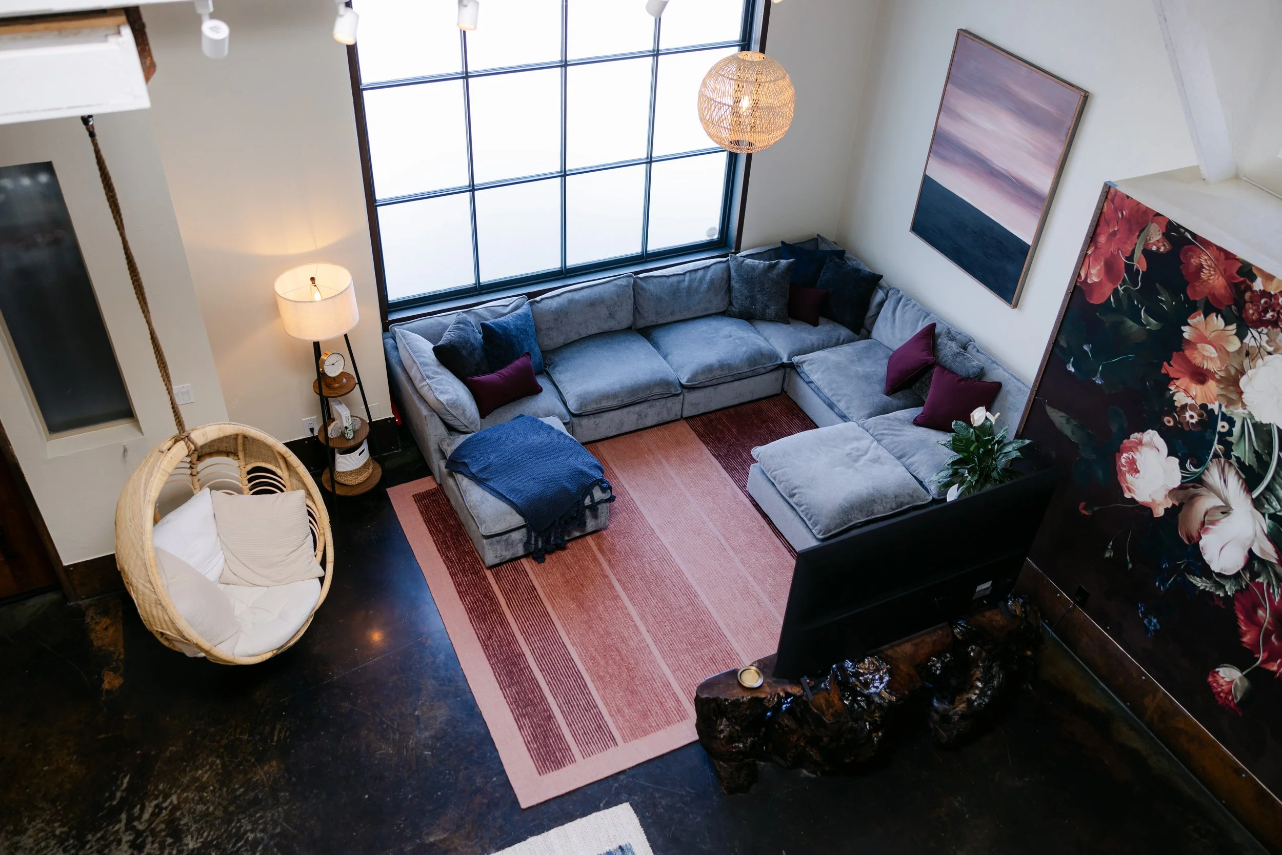 A cozy living room featuring a large gray sectional sofa with throw pillows in various dark colors, a pink and orange striped area rug, a woven hanging light fixture, a standing lamp with a white shade, a small round side table with a clock and decor