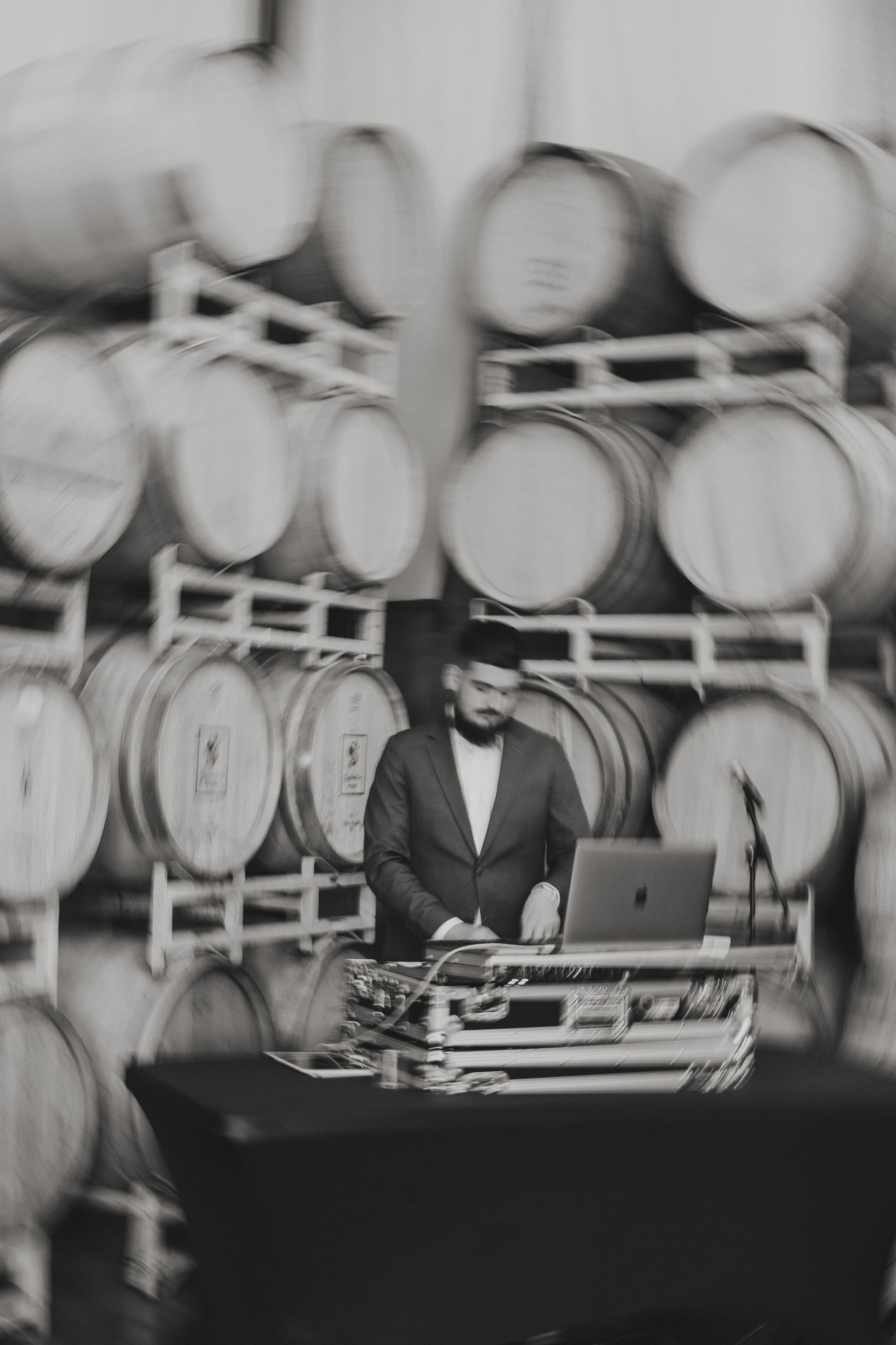Black and white photo of a man in a suit with glasses working on a laptop in front of stacked wine barrels in a winery storage room.