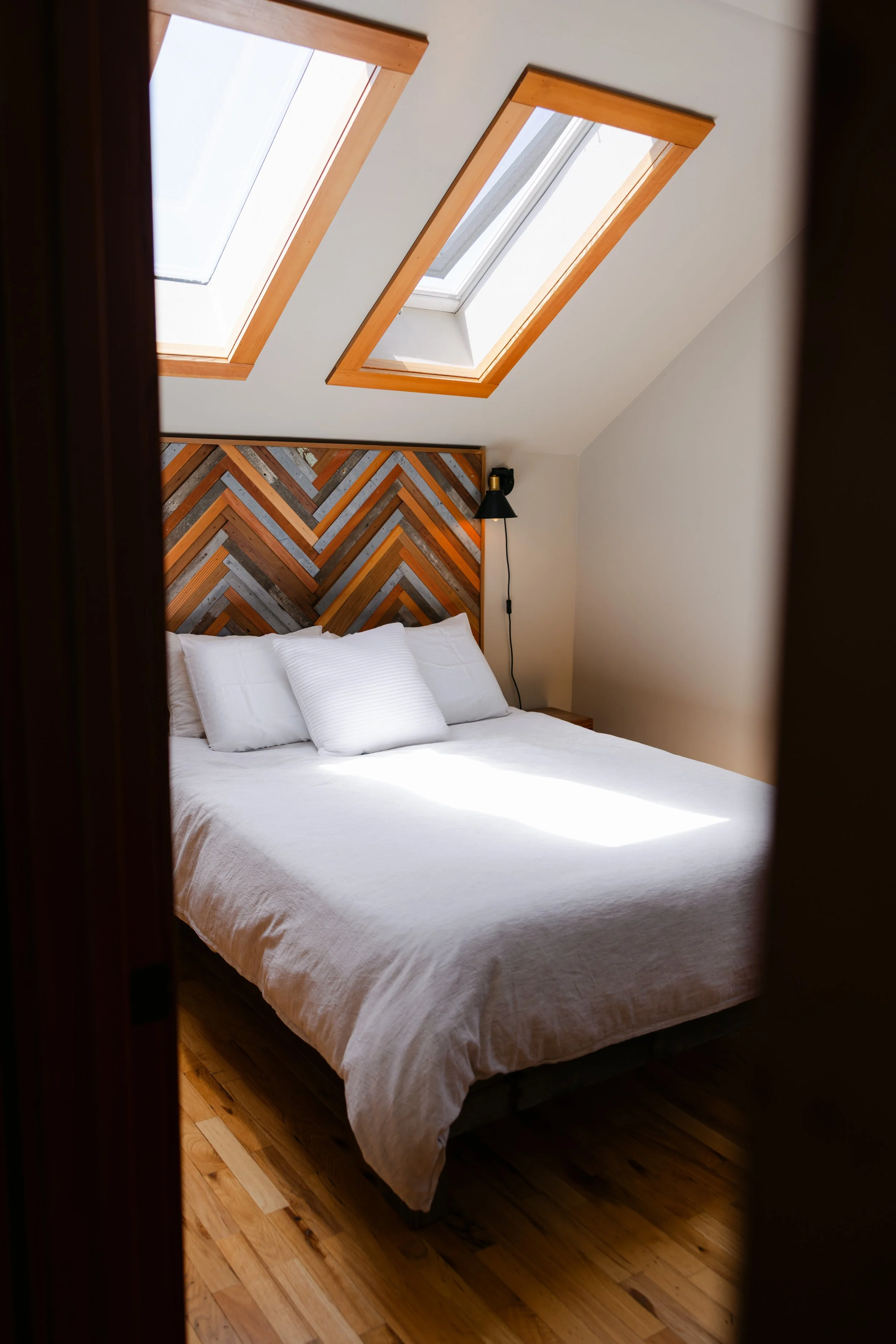 A bedroom with a white bed, pillows, wooden herringbone accent wall, and two skylight windows in the ceiling allowing natural light to enter.