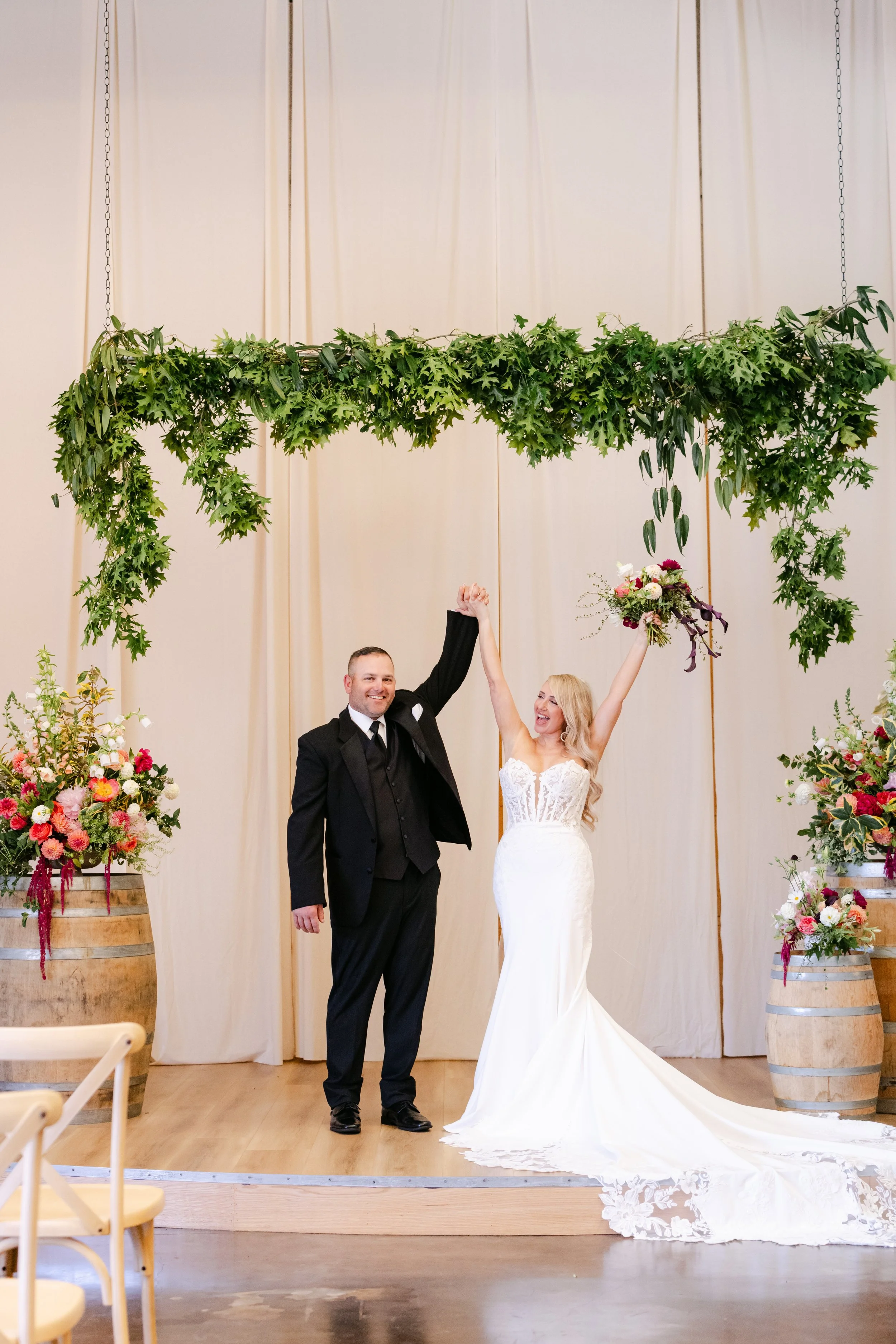 A wedding ceremony with a bride and groom celebrating. The bride is in a white lace dress and holding a bouquet, raising her arm, and the groom is in a black tuxedo. They are standing under a green leafy decoration hanging from the ceiling with small