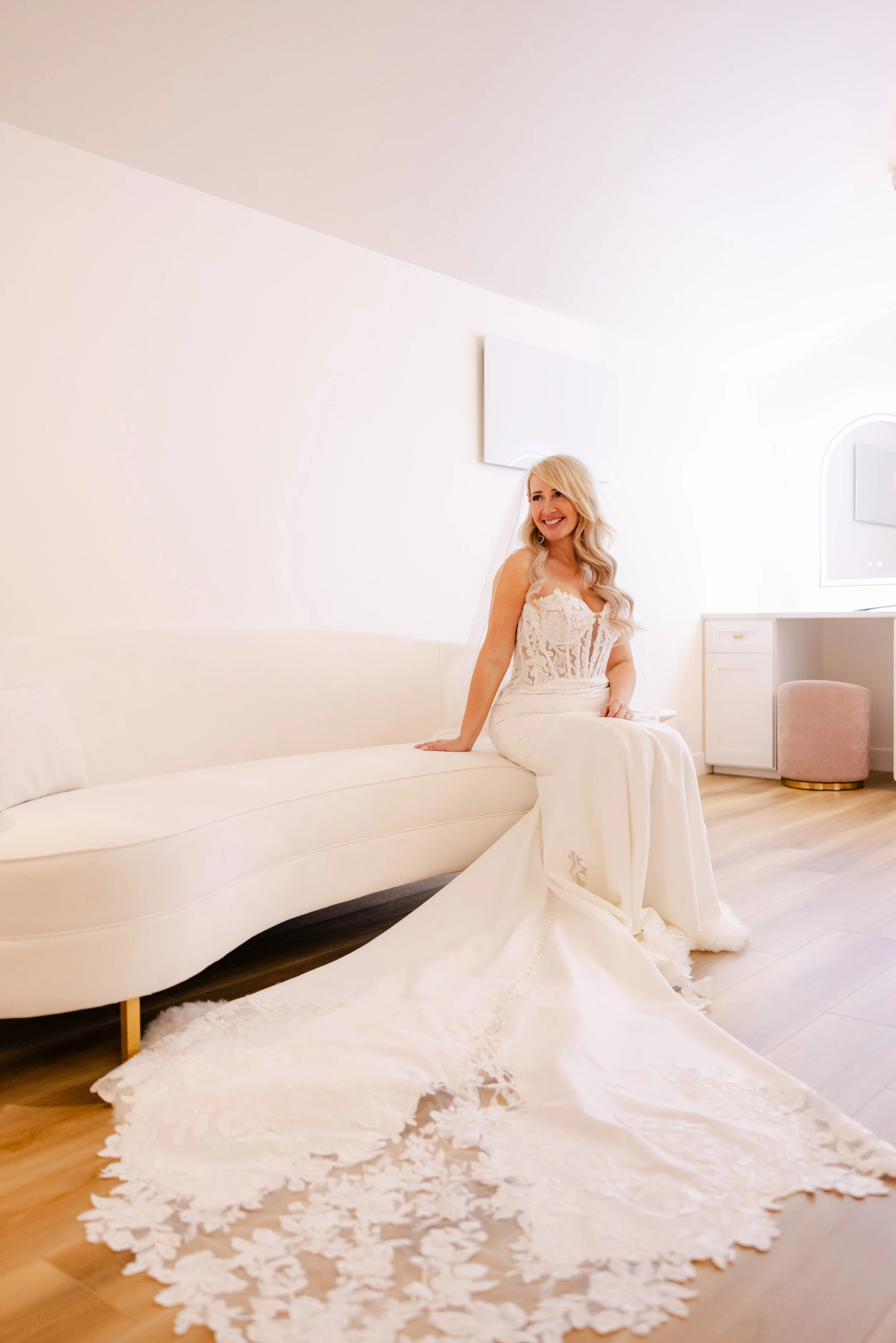 A woman in a white lace dress sitting on a white sofa in a bright room. The dress has a long train with floral patterns on the edge spread on the wooden floor.