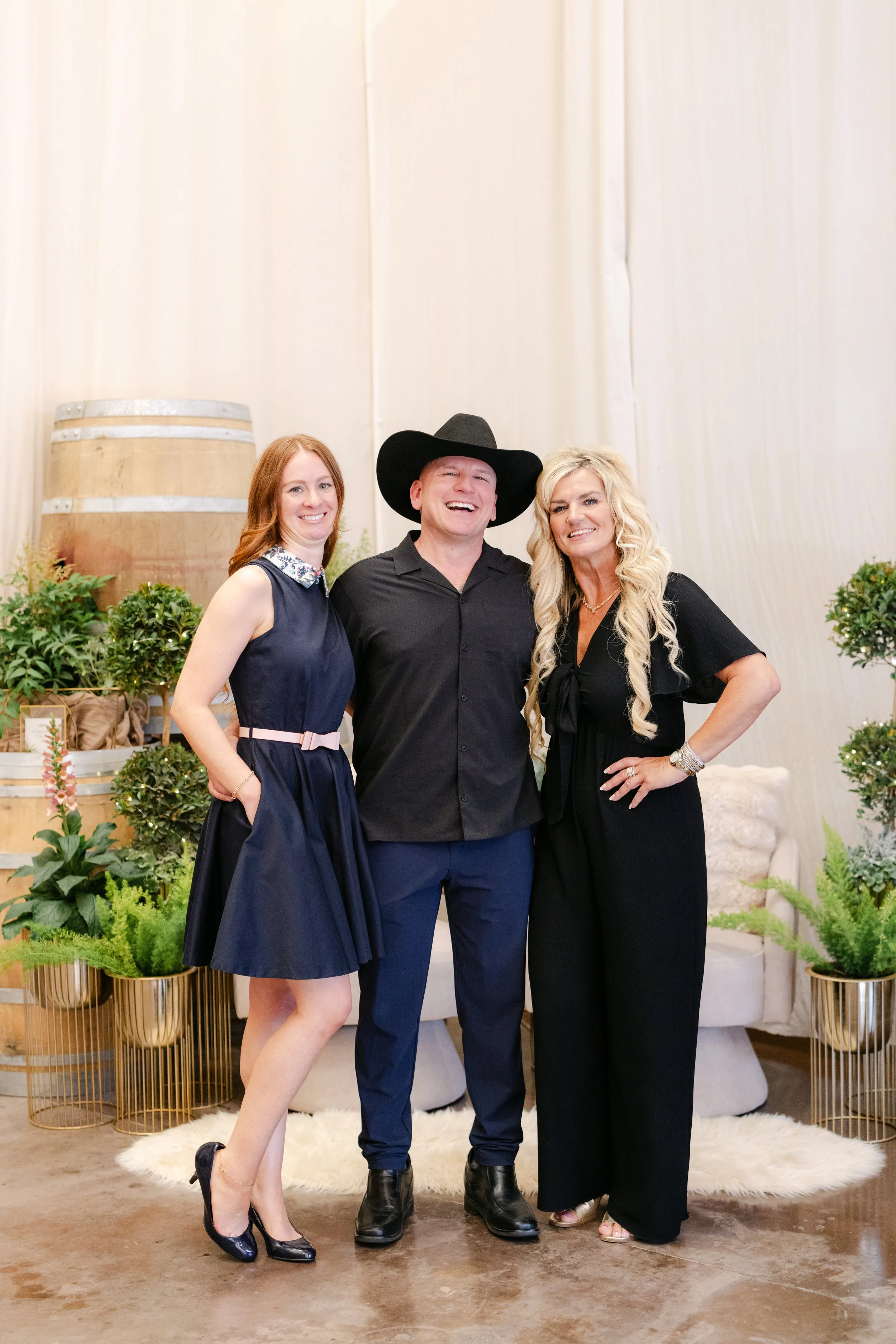 Three adults, two women and one man, are smiling and posing together indoors. The woman on the left has red hair and is wearing a navy blue dress with a floral collar and black heels. The man in the middle is wearing a black cowboy hat, black shirt, 