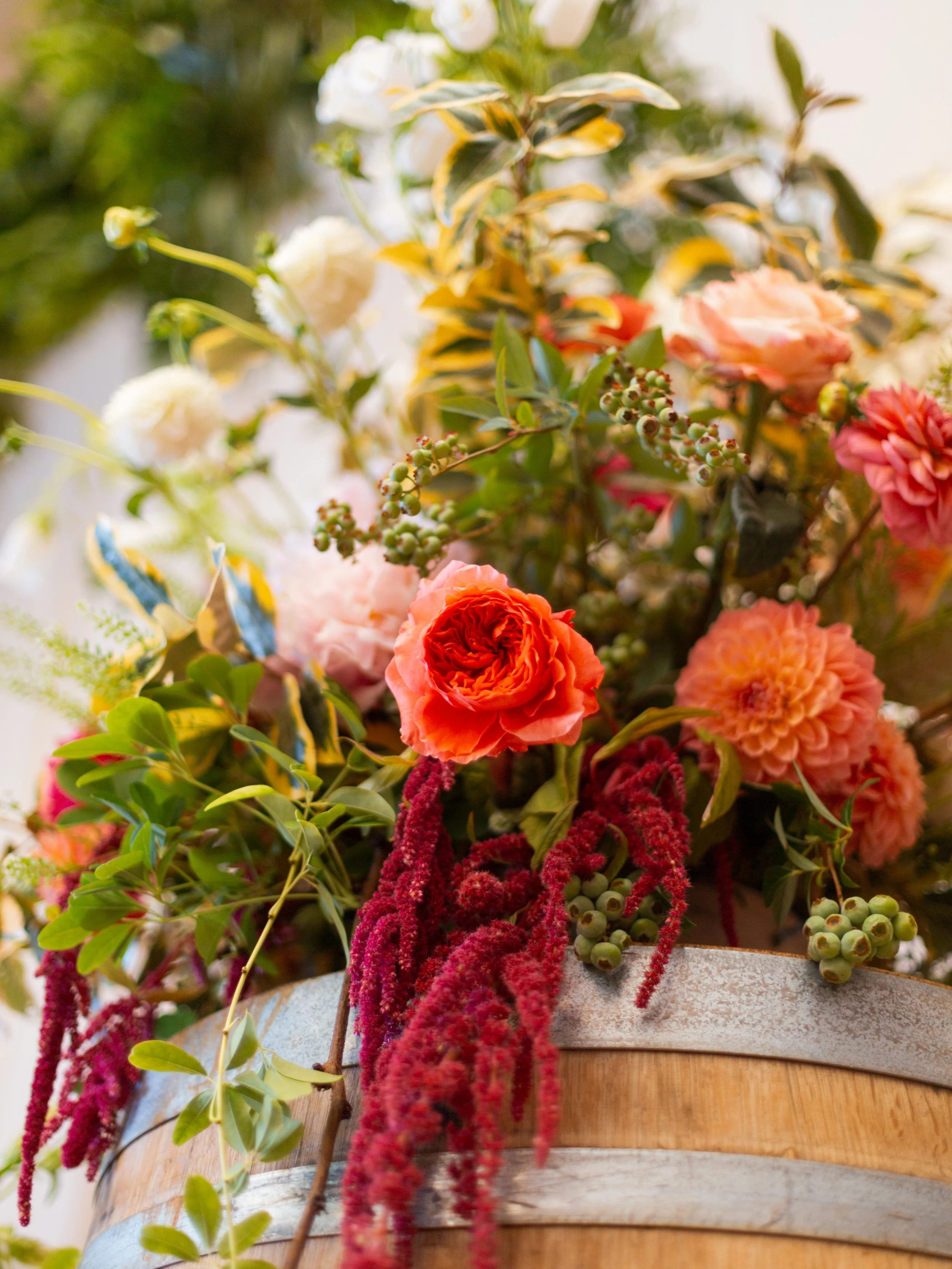 A close-up of a floral arrangement in a wooden container, featuring pink and peach roses, green berries, variegated leaves, and deep red hanging amaranthus.