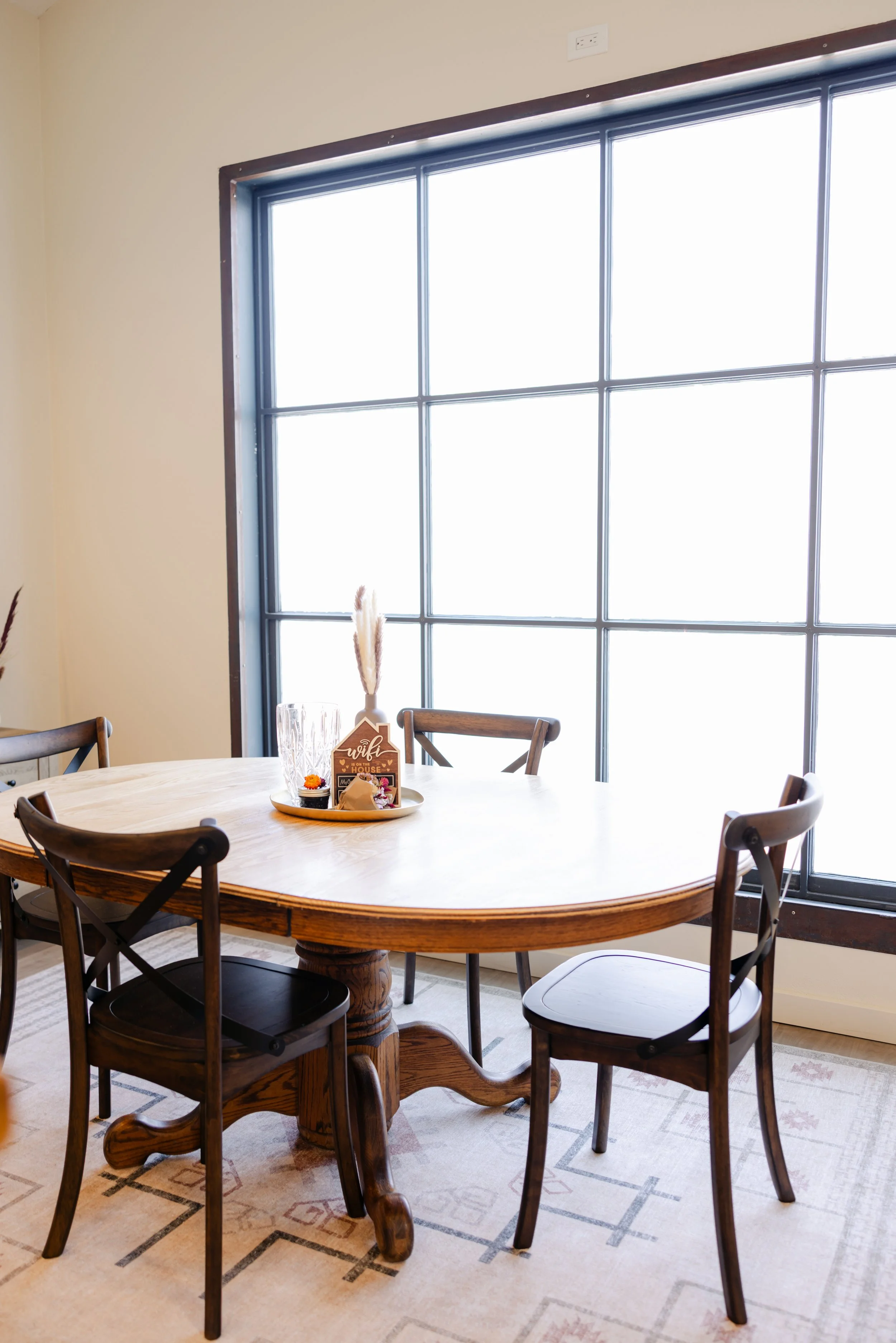 A round wooden dining table with six chairs, placed in front of a large window with black framing, with a decorative centerpiece and glassware on the table, and a beige patterned rug underneath.