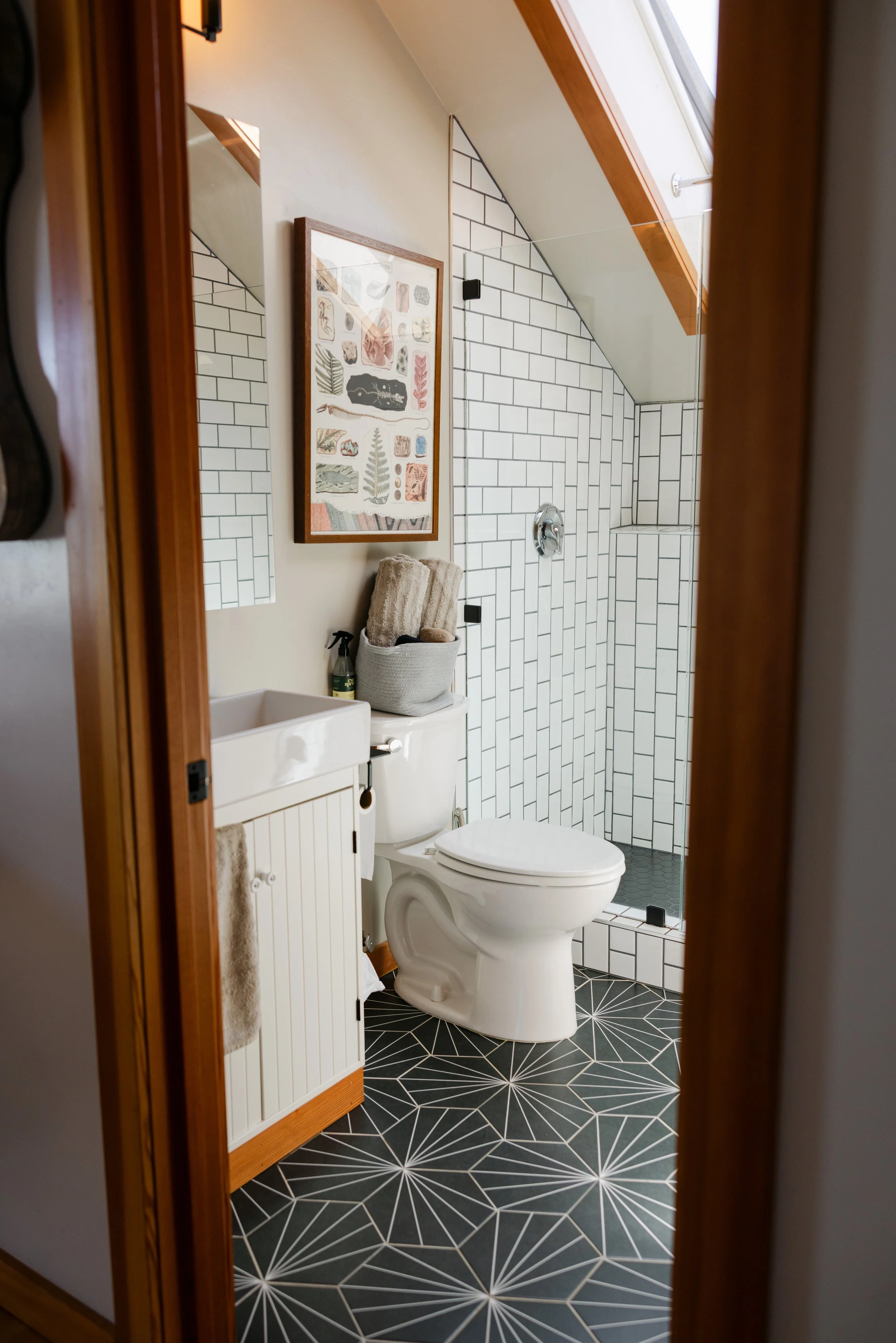 A bathroom with a white toilet, a small white cabinet, black geometric patterned floor, tiled walk-in shower with white subway tiles, a mirror on the wall, and a framed art print featuring plant and nature illustrations.