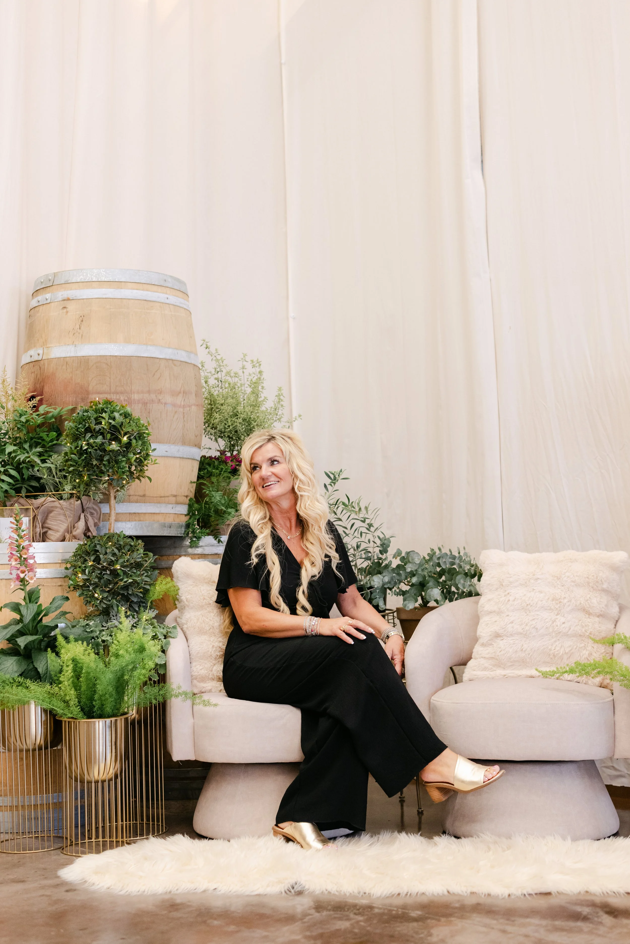 A woman with long blonde hair, wearing a black dress and gold shoes, sitting on a beige sofa surrounded by green plants and fur pillows in a cozy, well-decorated indoor space.
