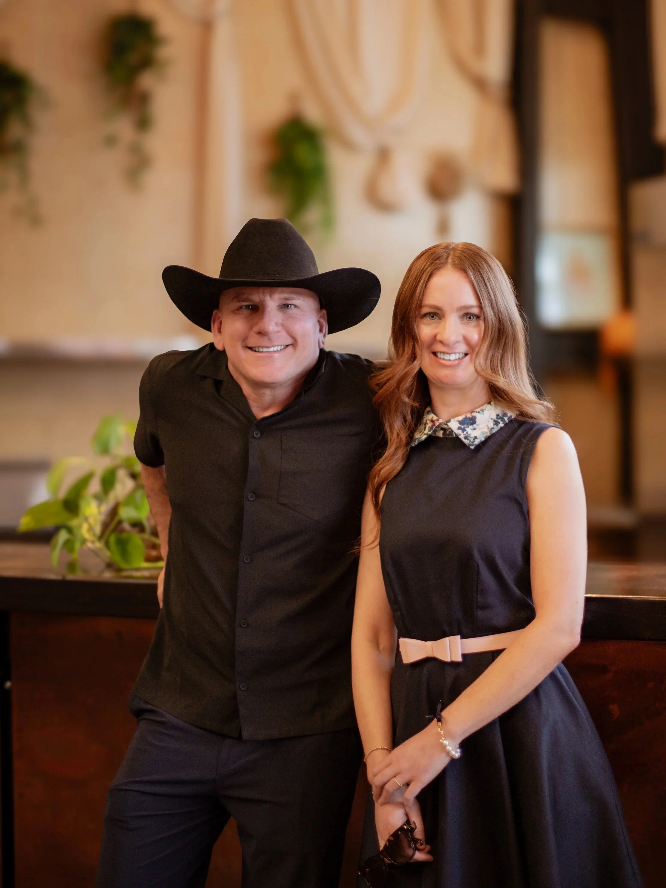 A man wearing a black cowboy hat and a black button-up shirt standing next to a woman with reddish-brown hair, wearing a dark dress with a floral collar and beige belt, in a warmly lit indoor setting.