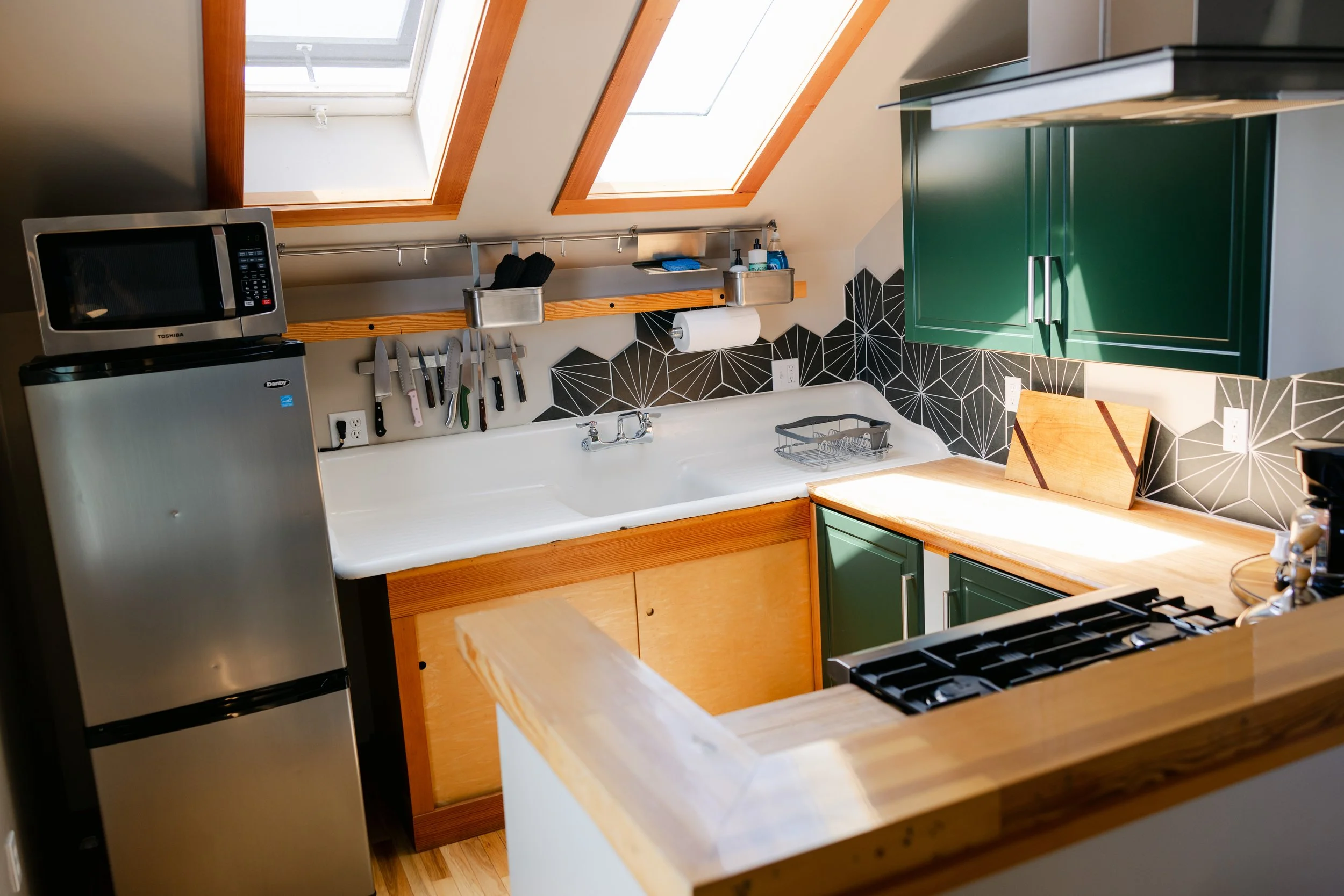 A kitchen with skylight windows, a microwave on top of a small refrigerator, a white sink, green cabinets, a black and white geometric tile backsplash, and kitchen tools hanging on a rail.