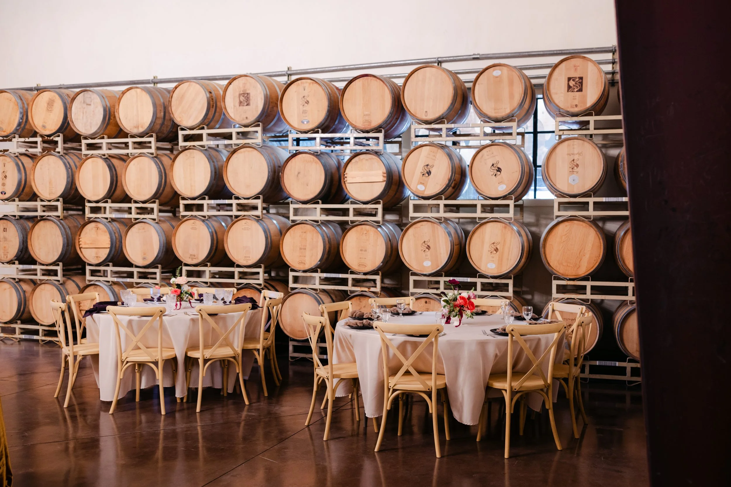 Rows of wine barrels stacked on racks in a winery, with tables and chairs set for dining in the foreground.