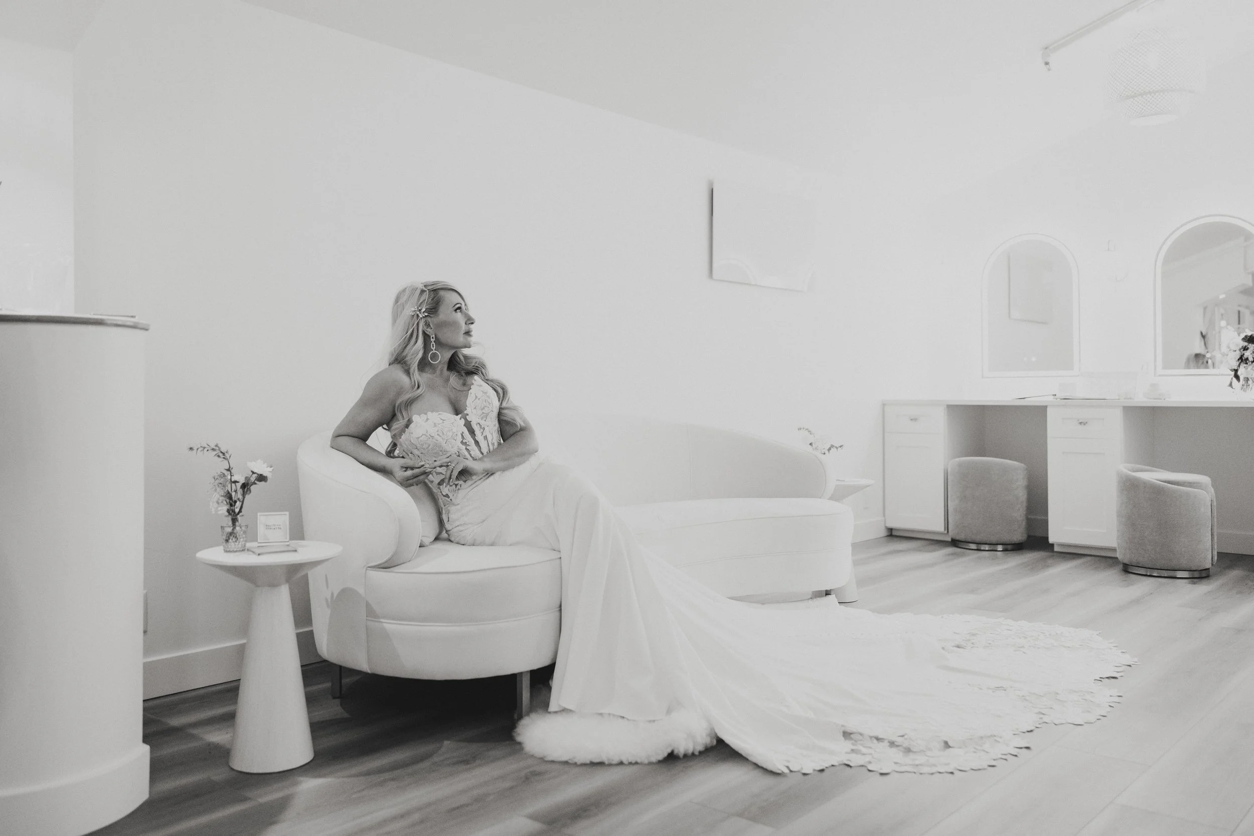 A woman in a wedding dress sitting on a white sofa in a minimalist, modern room with white walls and furniture, including a side table with flowers and a small sign.