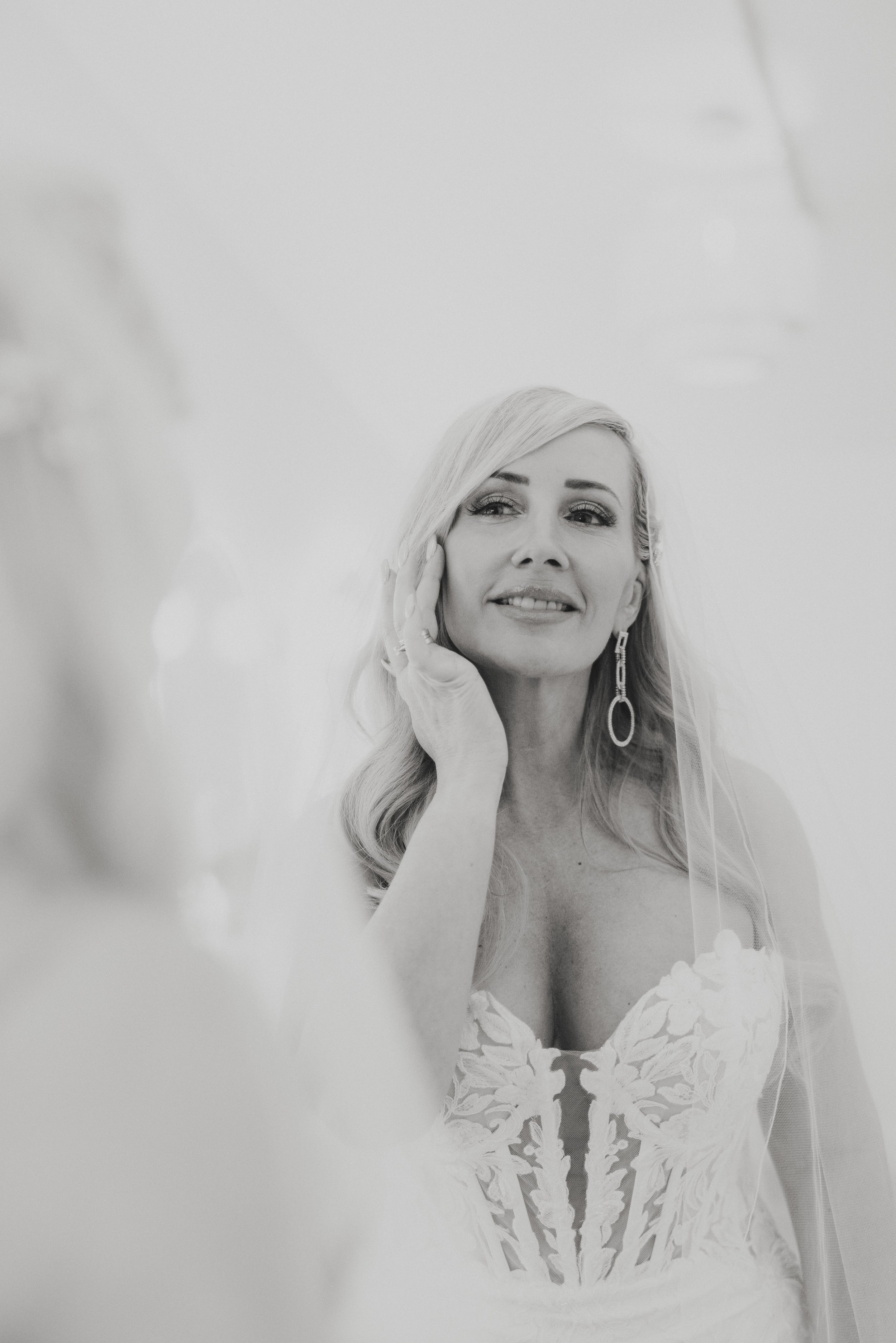 Black and white photo of a woman in a lace dress with a veil, touching her face, and smiling softly.