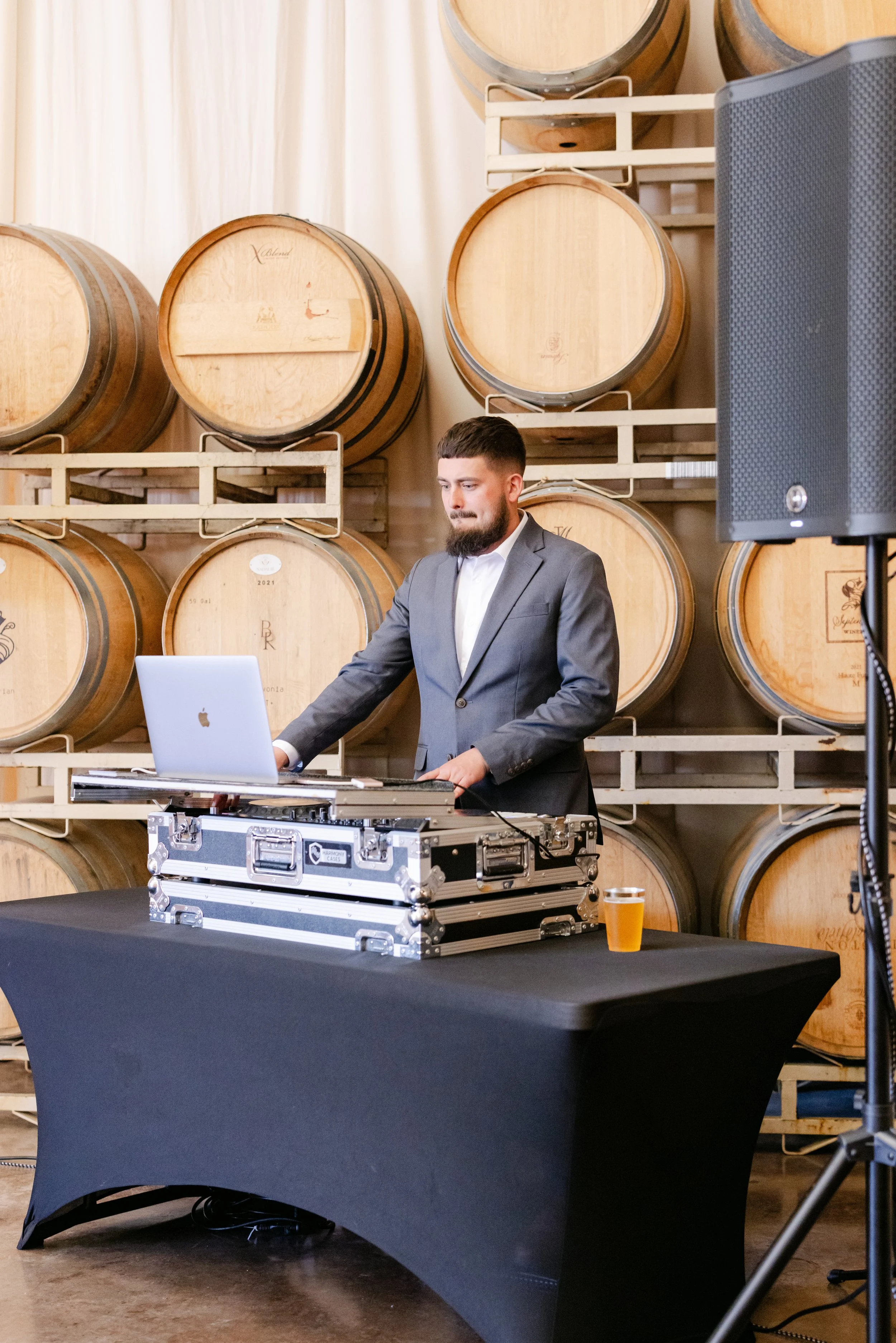 A man in a gray suit is DJing at an event with large wooden wine barrels in the background. He is operating DJ equipment with a laptop and a mixer, and there is a glass of beer on the table in front of him.
