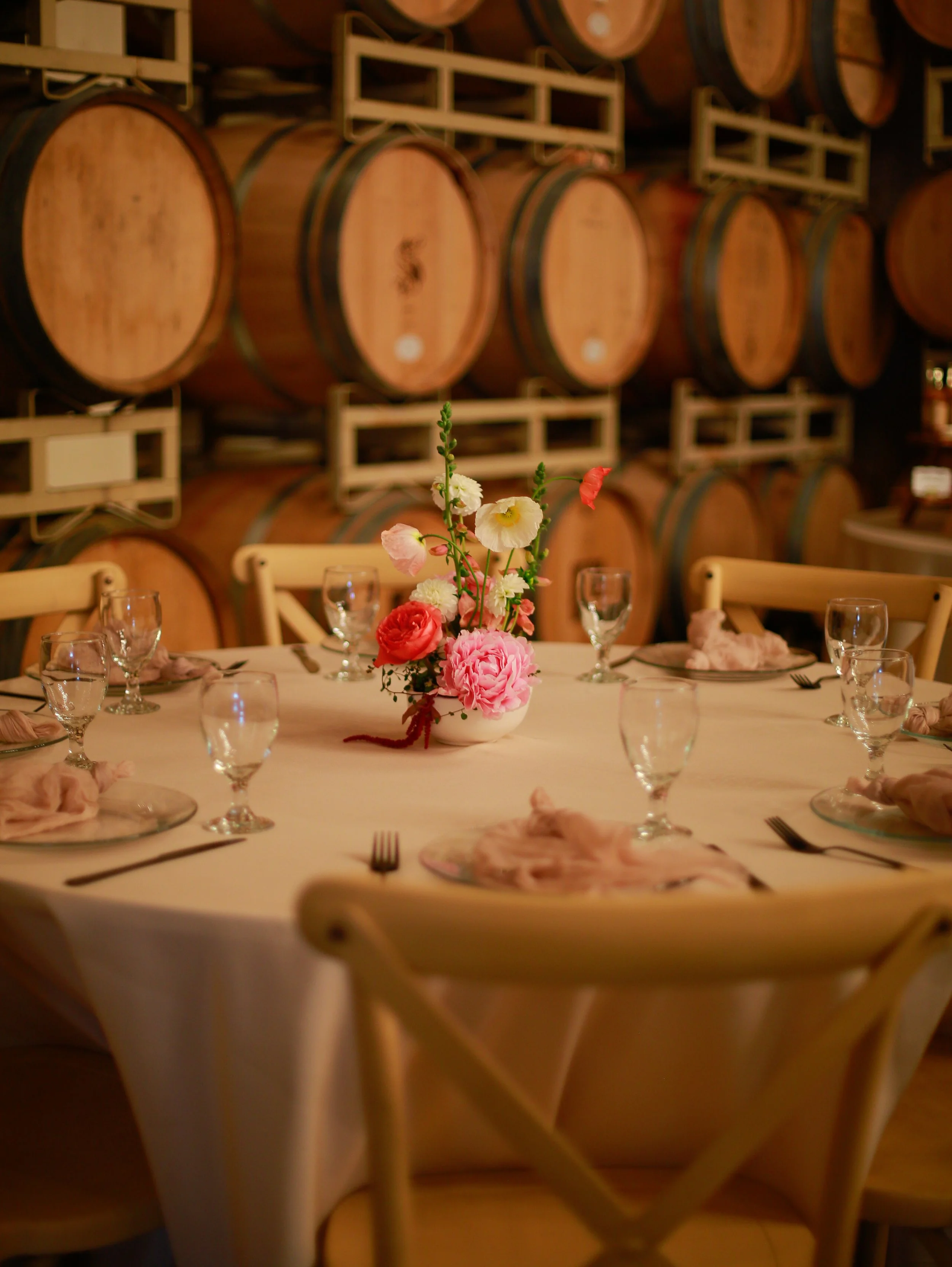 A round table set for a meal, decorated with a bouquet of pink, white, and red flowers in a white vase, surrounded by several wine glasses, plates with napkins, and silverware. In the background, there are large wooden barrels stacked on shelves.