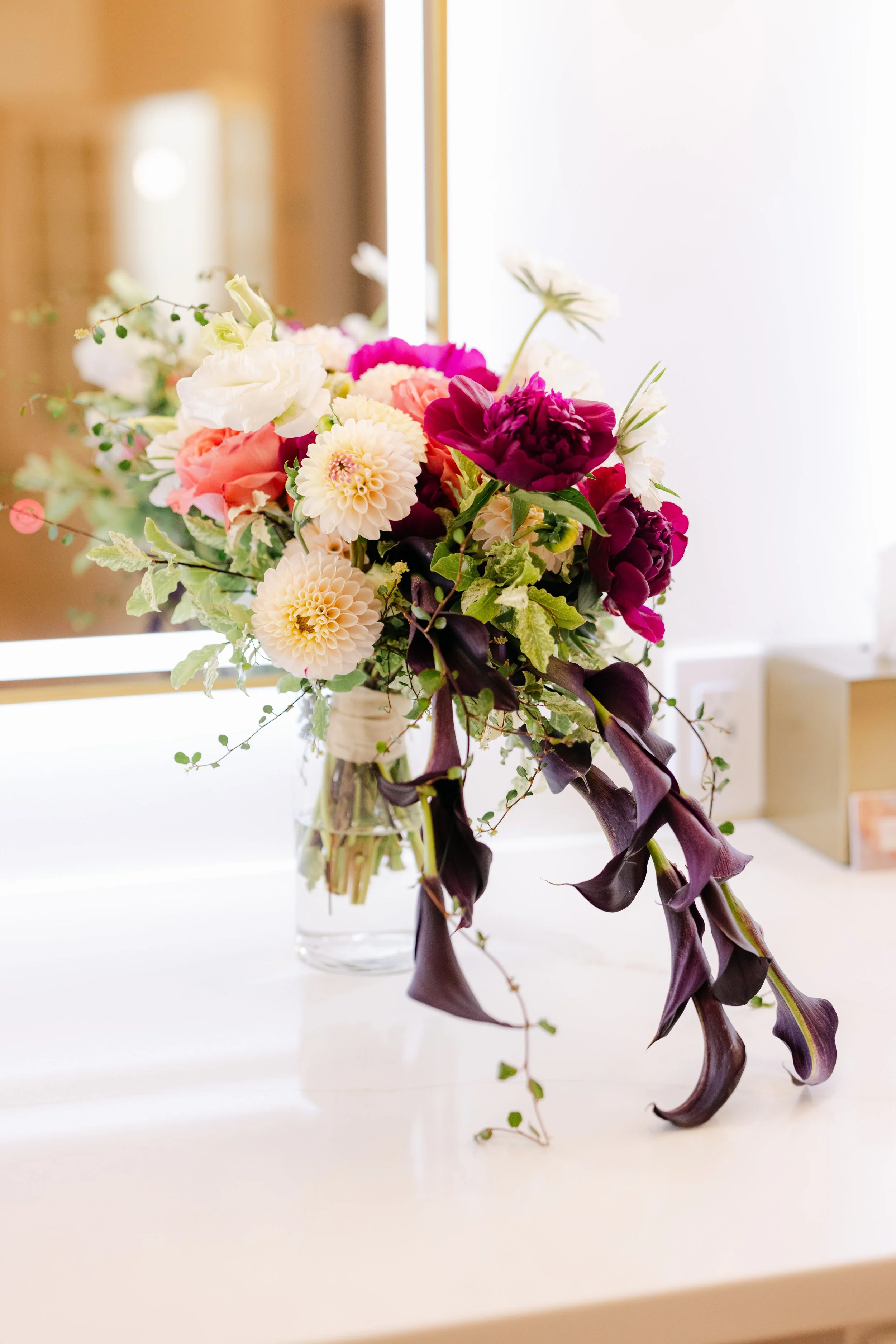 A colorful bouquet of flowers in a glass vase placed on a white surface, with a mirror and a wooden background.