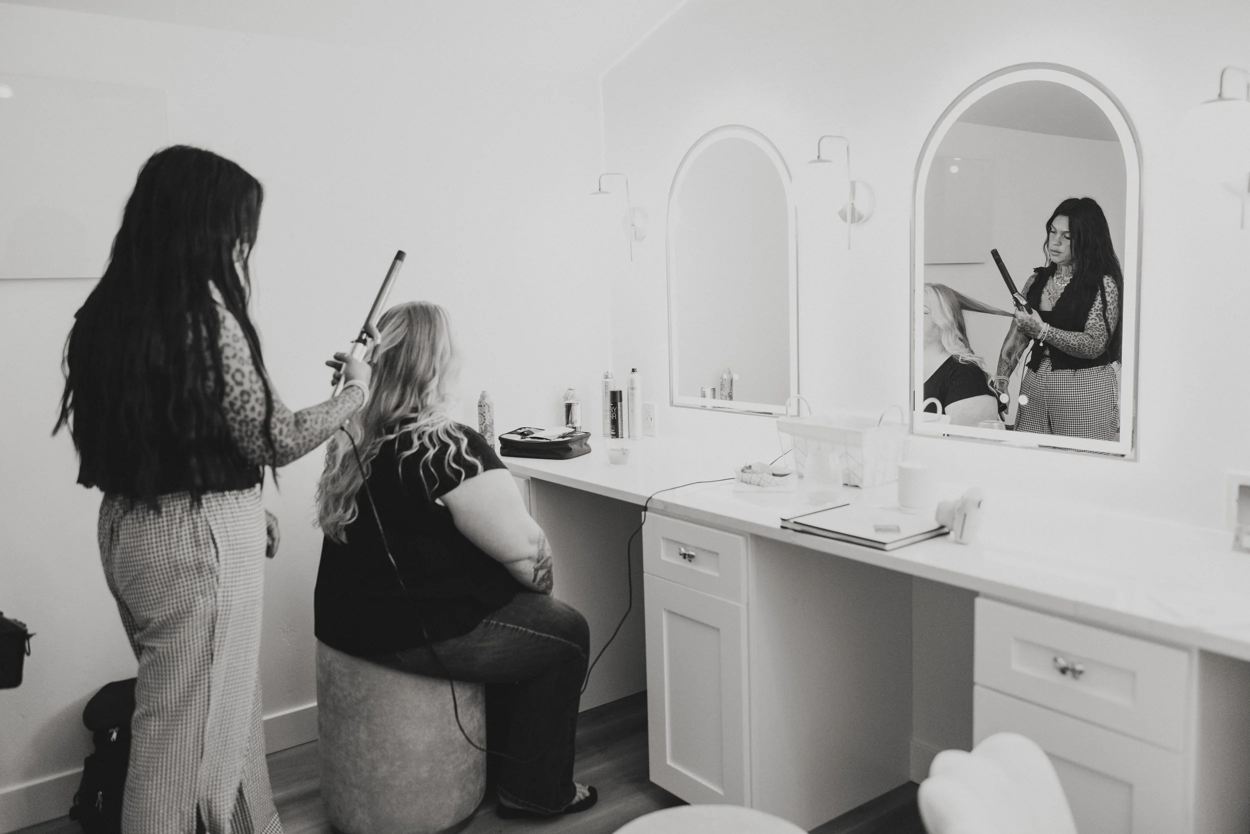A woman with long dark hair using a curling iron on another woman with blonde hair seated in a salon chair. A mirror reflects the scene, and various hair products are on the counter.