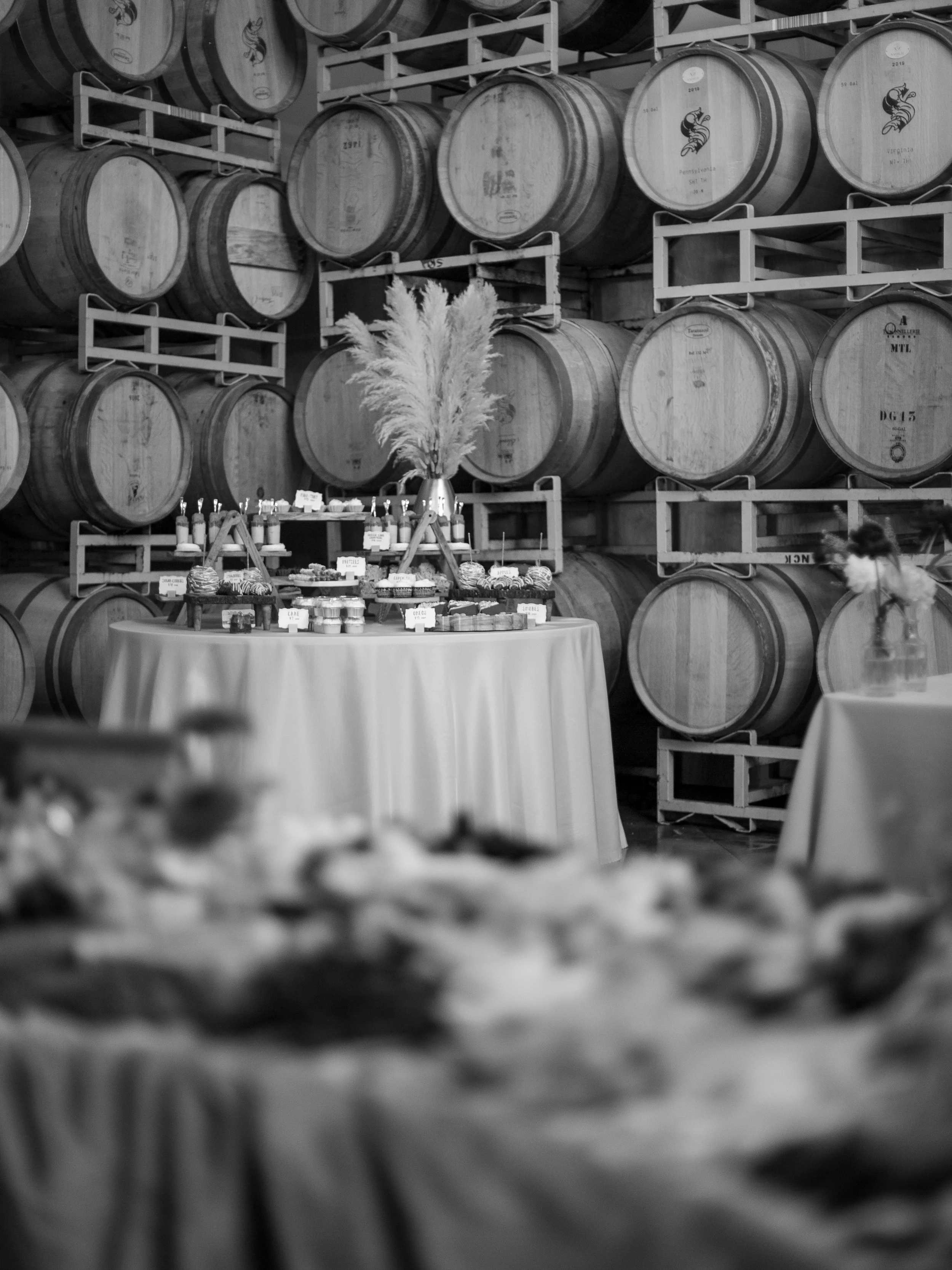Black and white photo of a wine cellar with numerous stacked barrels on racks. In front, there is a table with various desserts and a vase with pampas grass.