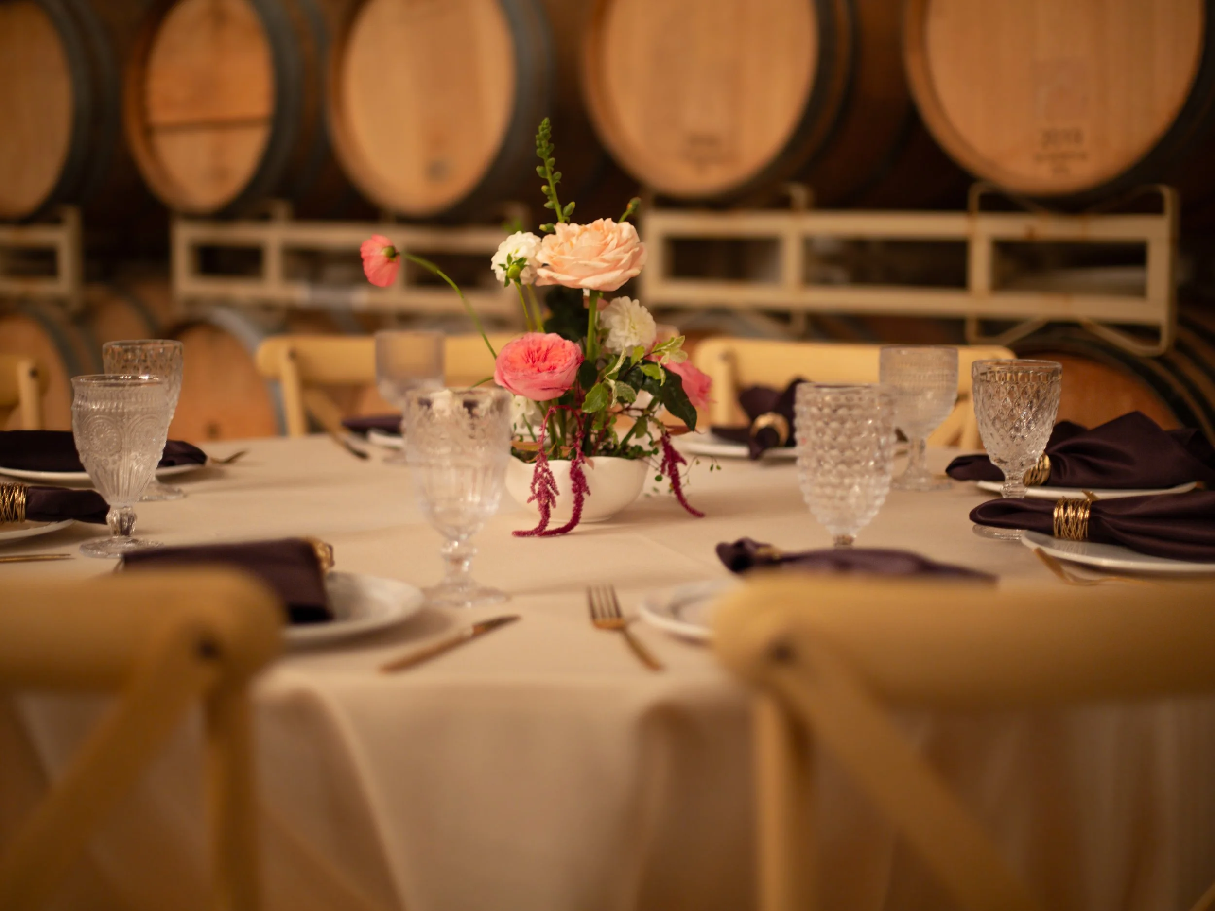 A floral centerpiece with pink, white, and peach flowers on a table set with glasses and embroidered napkins, in a room with wine barrels in the background.