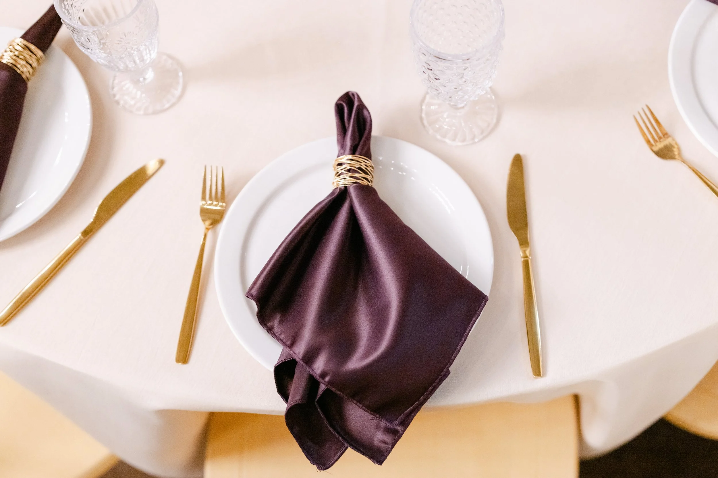 Table setting with a white plate, gold flatware, a folded purple napkin wrapped with gold wire, and crystal glasses on a white tablecloth.