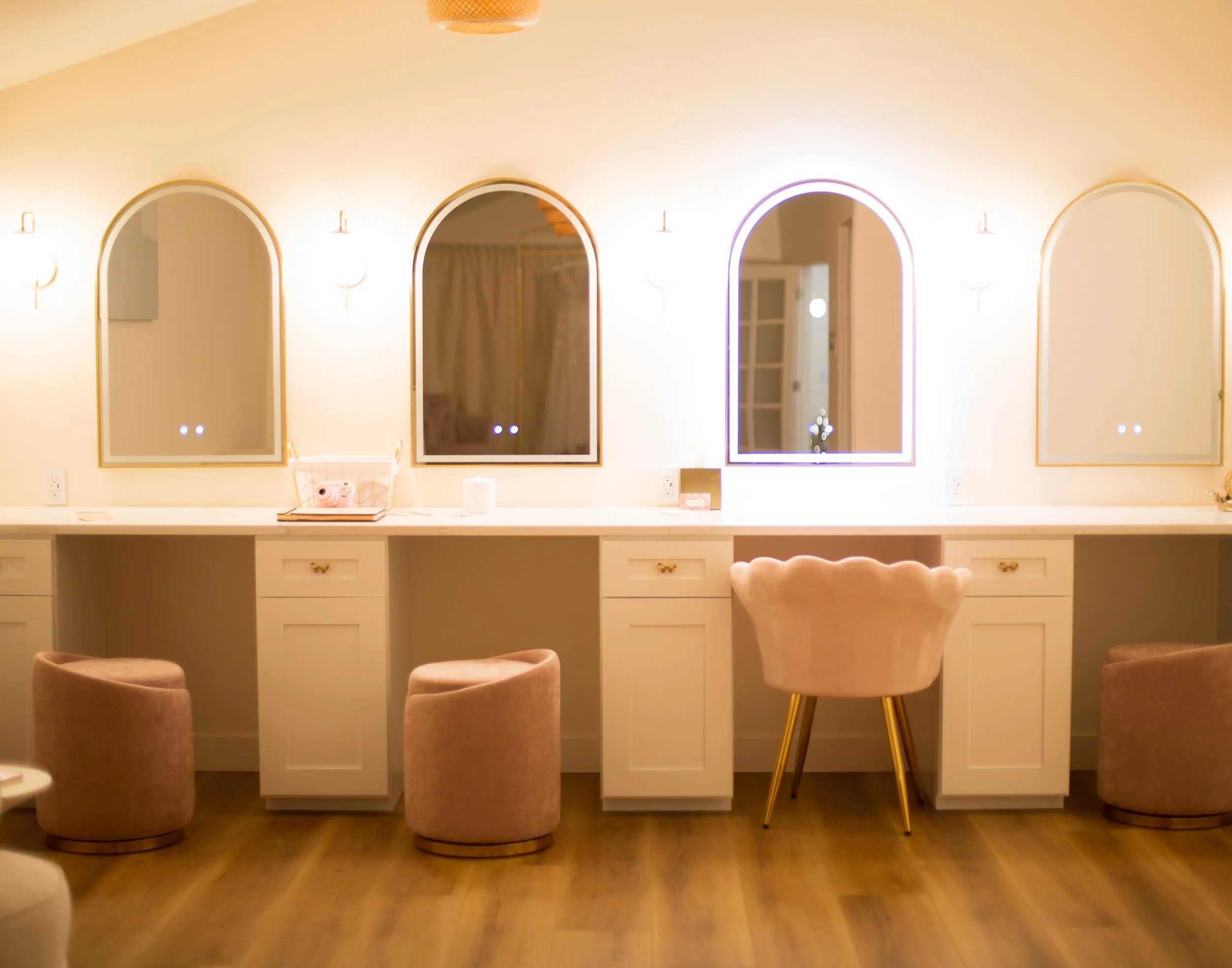 Interior of a modern salon or barbershop with a white counter with four drawers, three upholstered chairs in front, and three mirrors with rounded tops trimmed with gold, the wall behind has soft lighting fixtures.