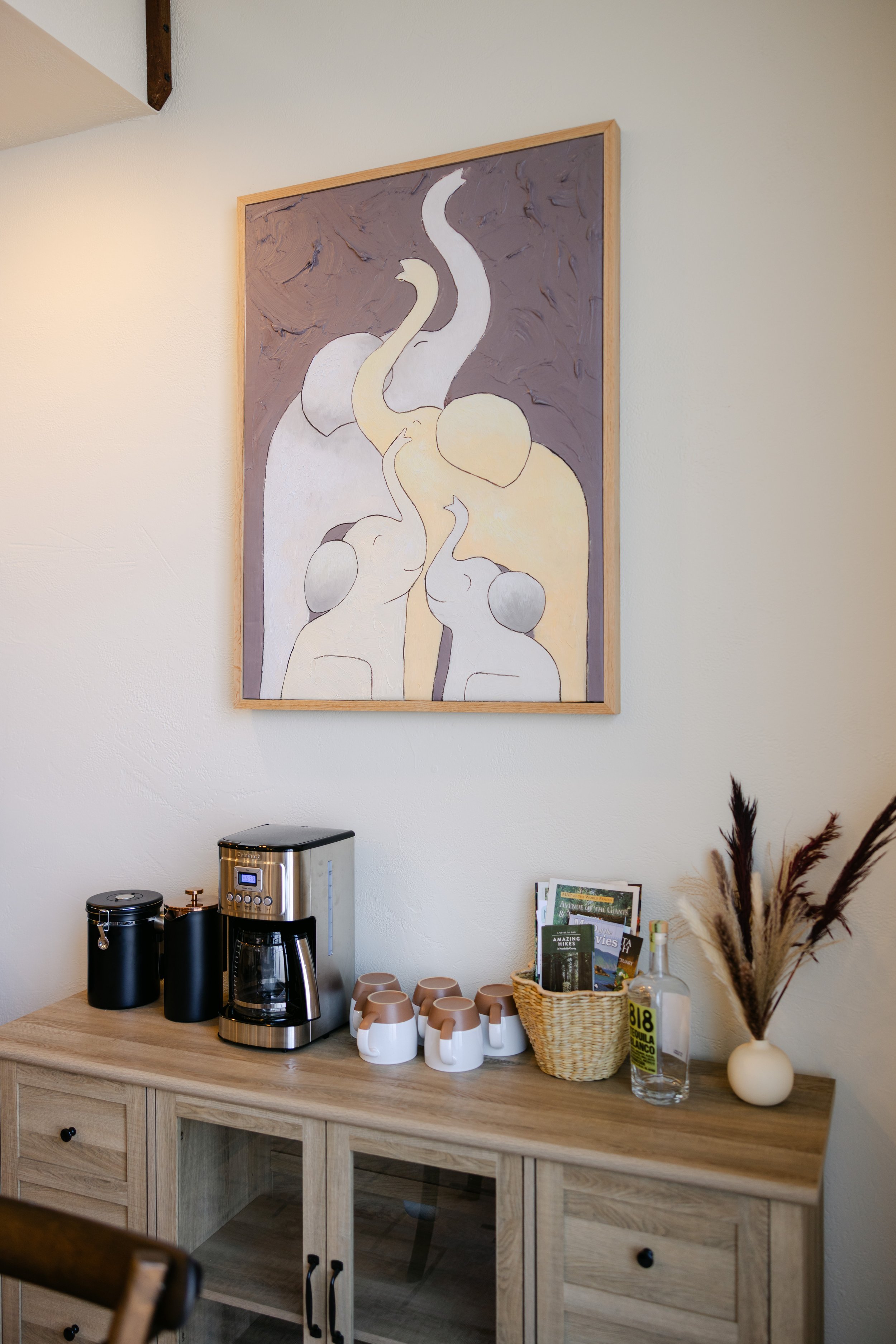 Wooden cabinet with black knobs, coffee-making items including a coffee machine and drip coffee maker, ceramic mugs with brown handles, decorative items, brochures, a glass bottle with pampas grass, and a framed abstract artwork on the wall.