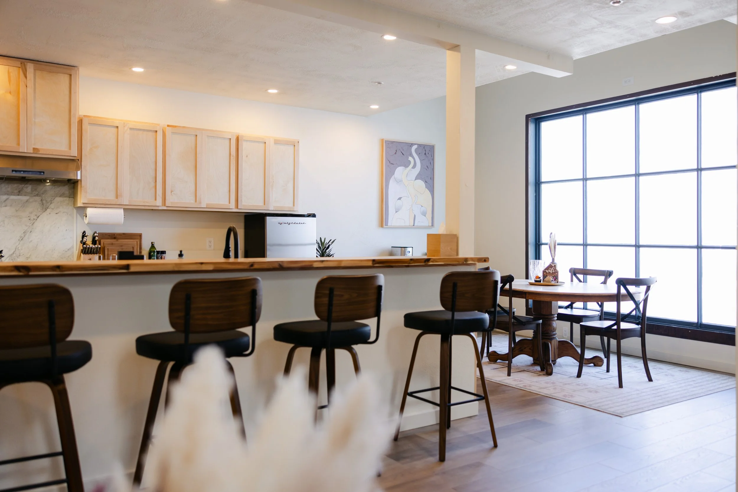 A modern kitchen and dining area with a row of black and wooden chairs at a white kitchen island, large windows allowing natural light, and light-colored wooden cabinets.