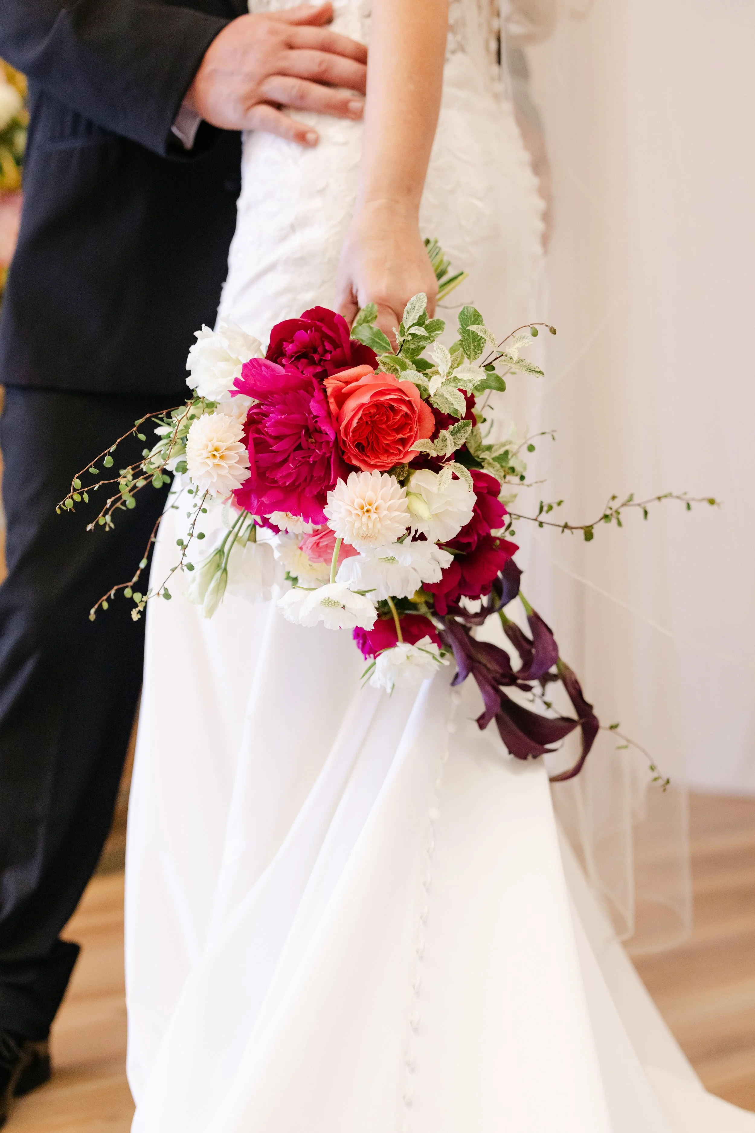 Close-up of a bride holding a colorful bouquet of flowers, with a groom standing beside her. The bride's hand is visible, and part of her white wedding dress is shown. The groom is wearing a dark suit.