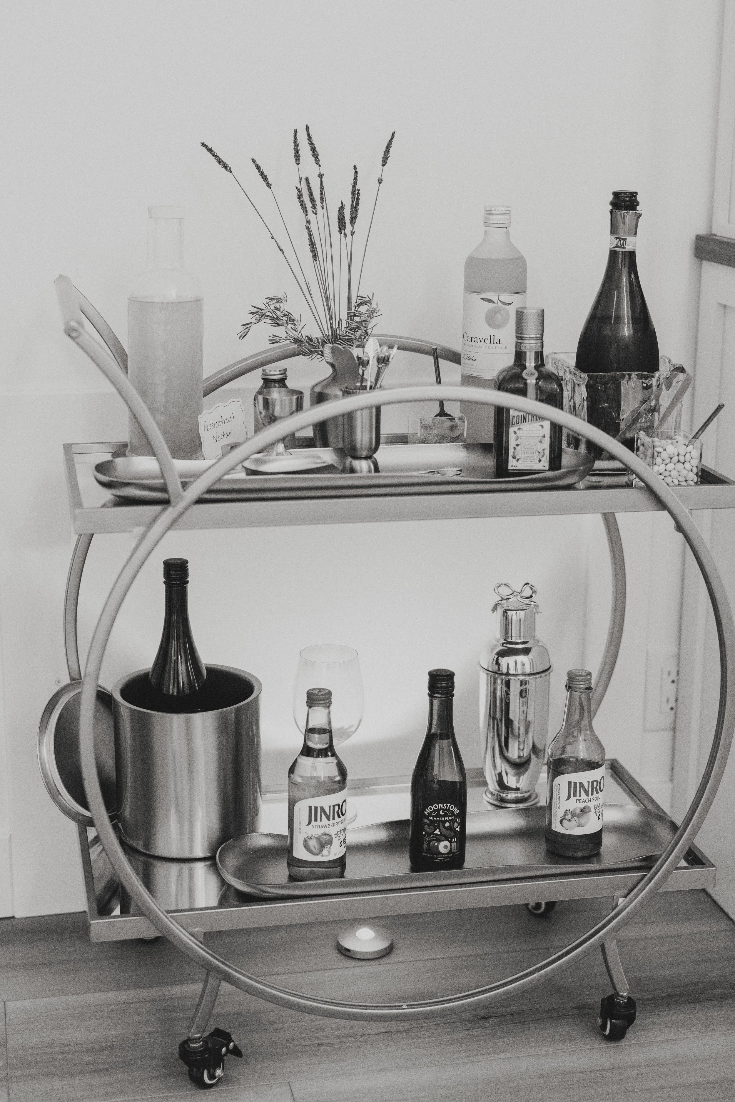A bar cart with various bottles of alcohol, including Jinro peach soju, Moonstone summer plum, and Courvoisier cognac, along with a metal ice bucket, a cocktail shaker, a vase of dried lavender, and a small bowl of candy, positioned against a plain w