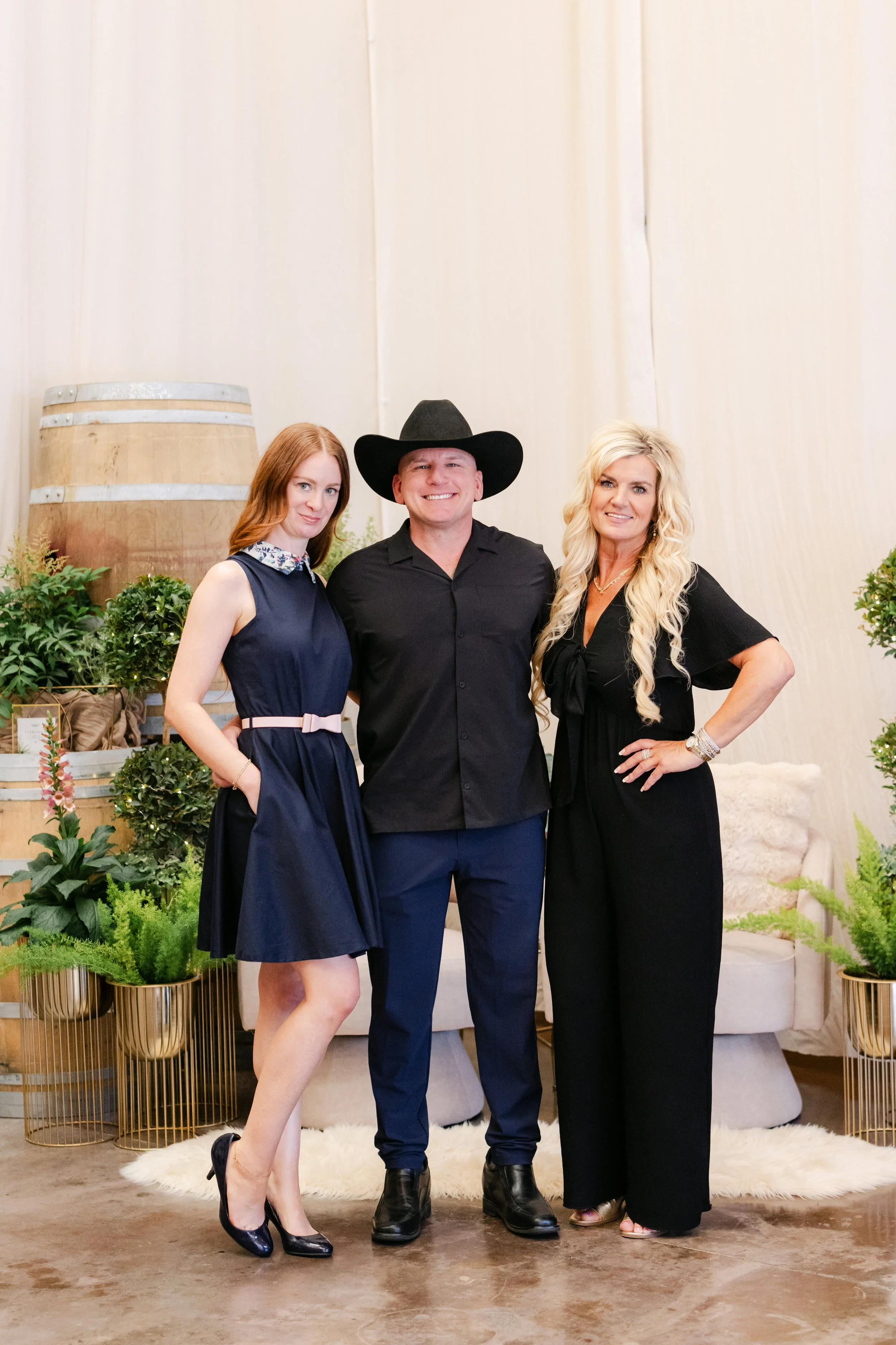 Three people standing together in a room decorated with plants and a large barrel in the background. The woman on the left has red hair and is wearing a navy blue dress, the man in the middle is smiling and wearing a black cowboy hat and black shirt,