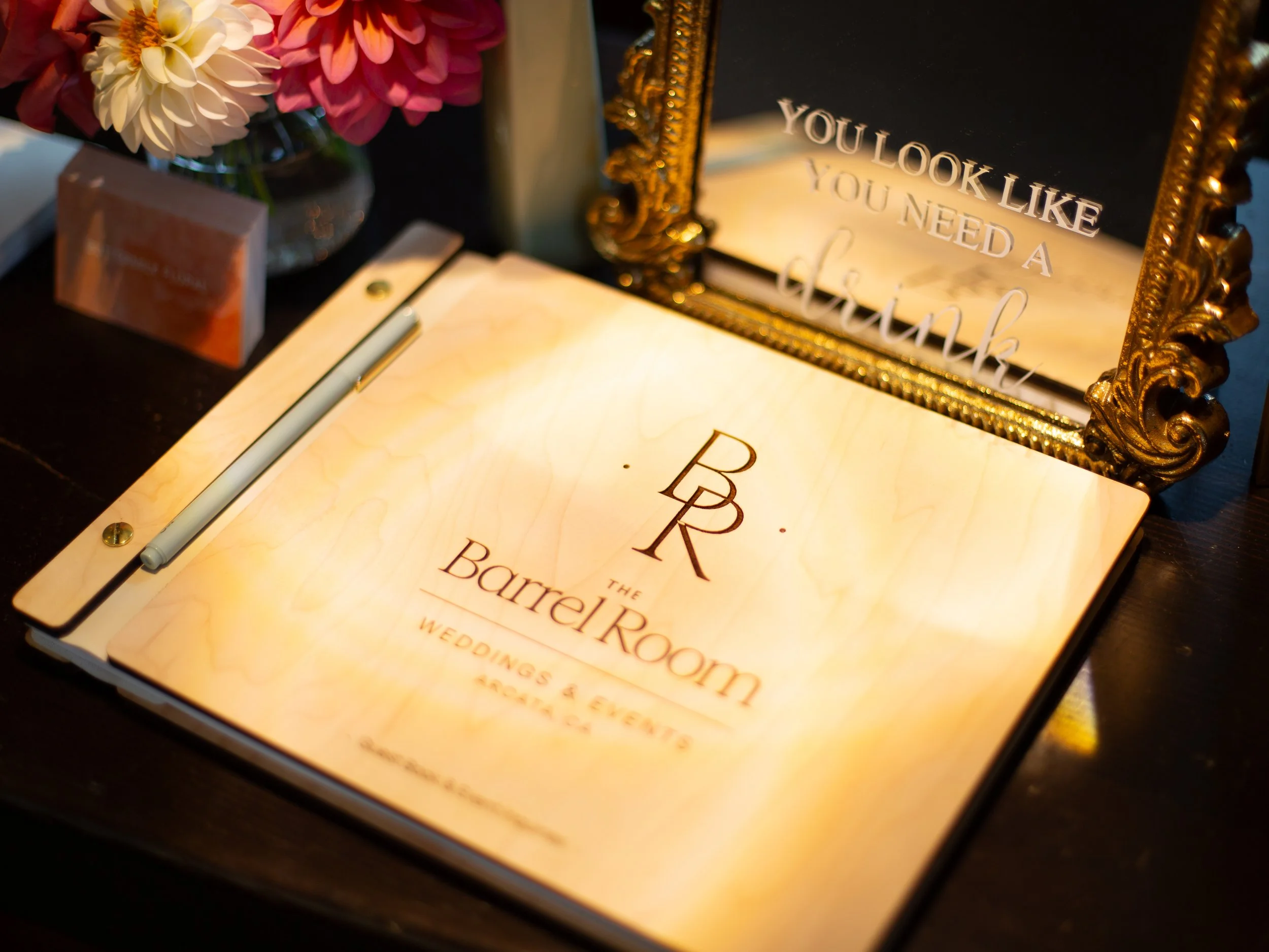 A decorative table setup with a floral arrangement, a sign with the phrase 'You look like you need a cocktail', a menu, a pen, and a small sign on a black surface.