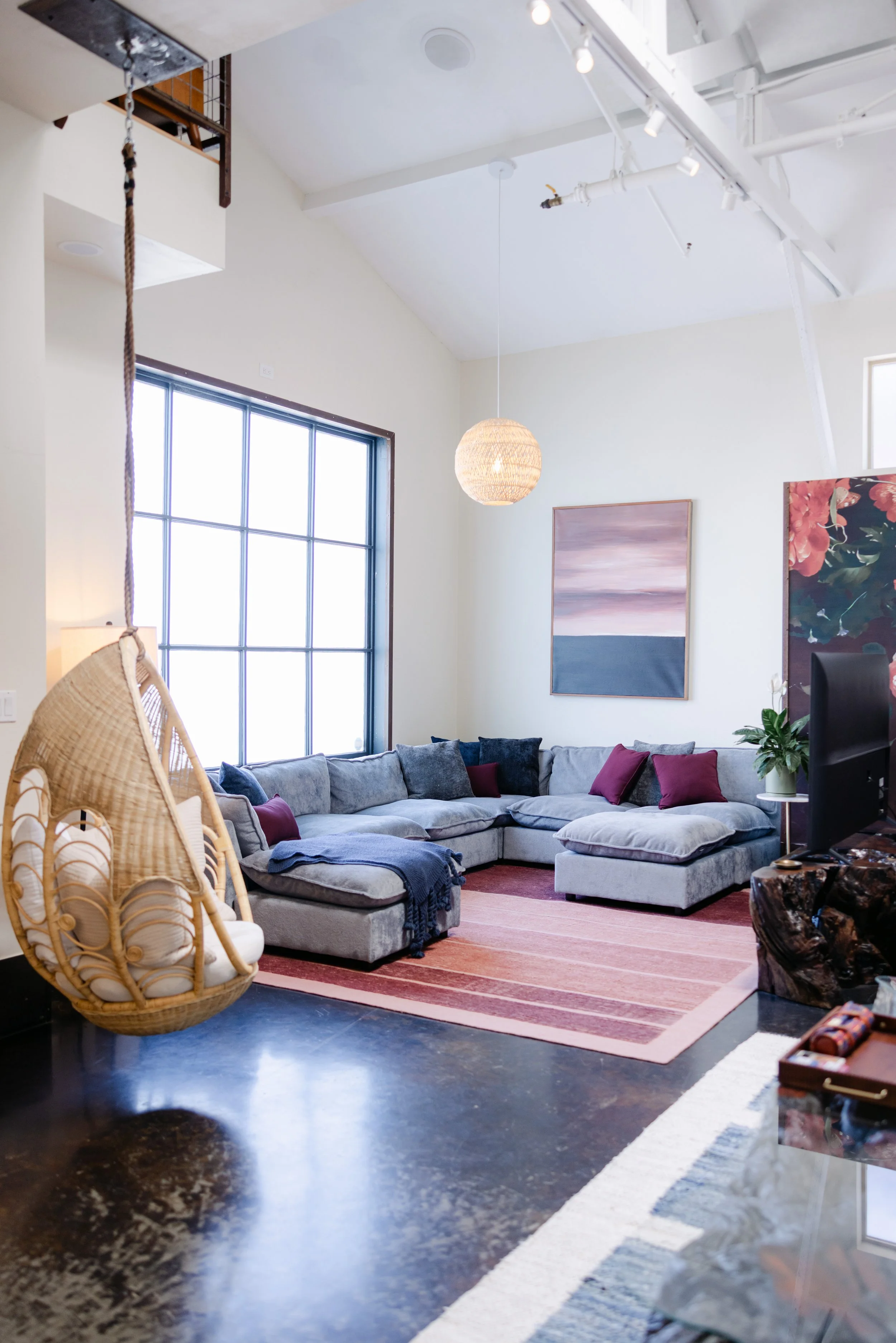 Living room with large sectional sofa, art paintings on wall, hanging pendant light, large window, and various decorative items like plant and stool.