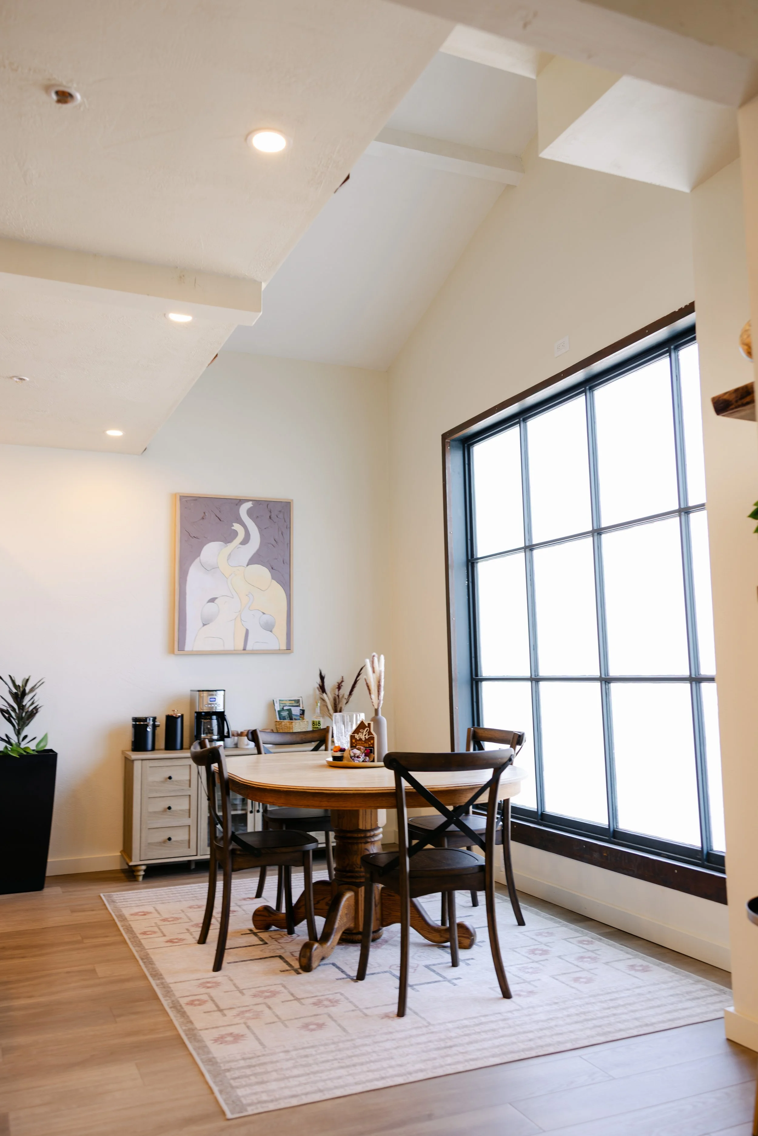 A cozy dining area with a round wooden table, four chairs, a white dresser with coffee-making supplies, a framed abstract artwork on the wall, and a large window with black framing allowing natural light. The room has light-colored walls, a beige are