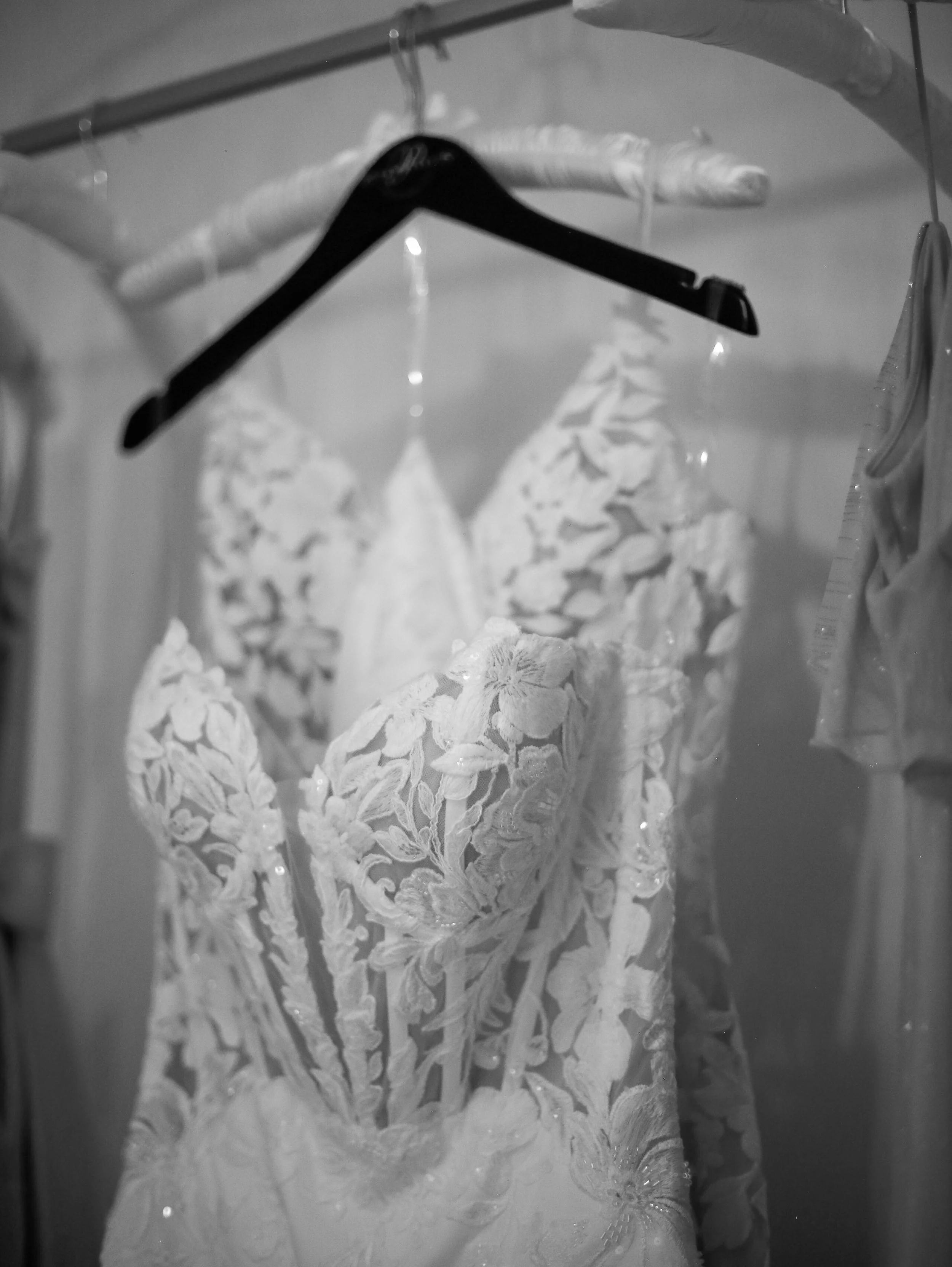 A close-up of a white lace wedding dress hanging on a black hanger, inside a closet with other clothing items visible.
