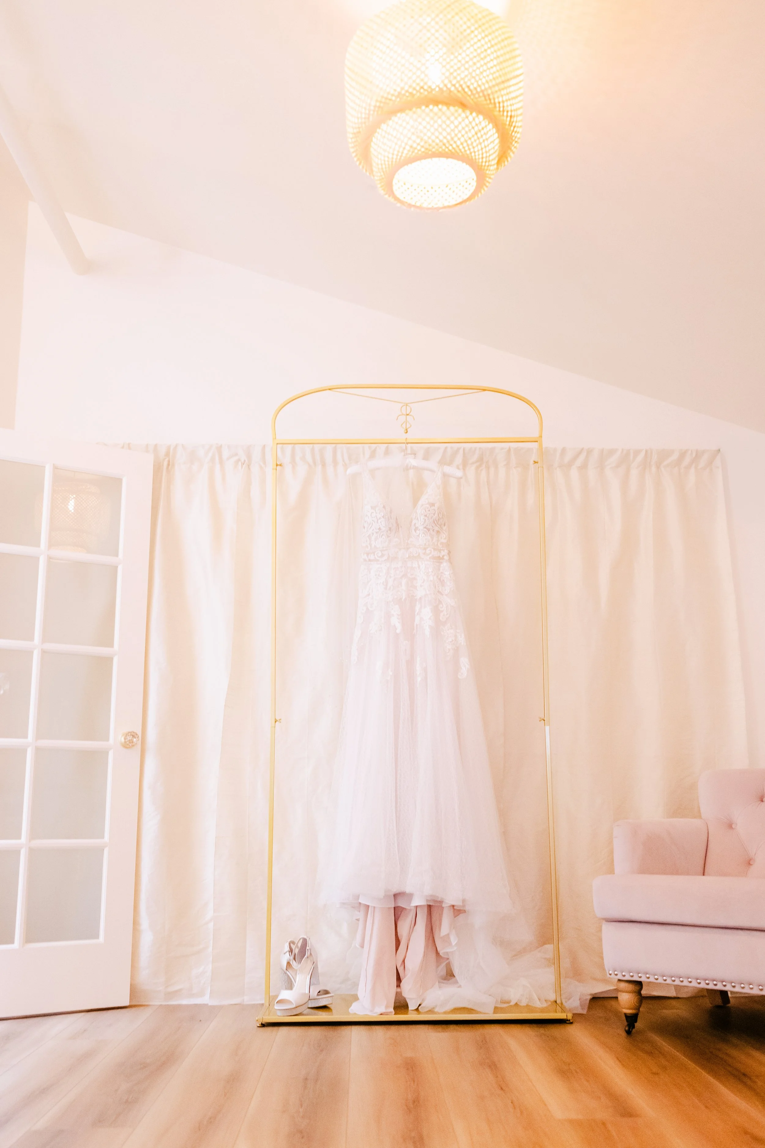 A wedding dress hanging on a gold frame, white curtains behind it, with a pair of white heels on the floor, a pink chair to the right, and an overhead light fixture.