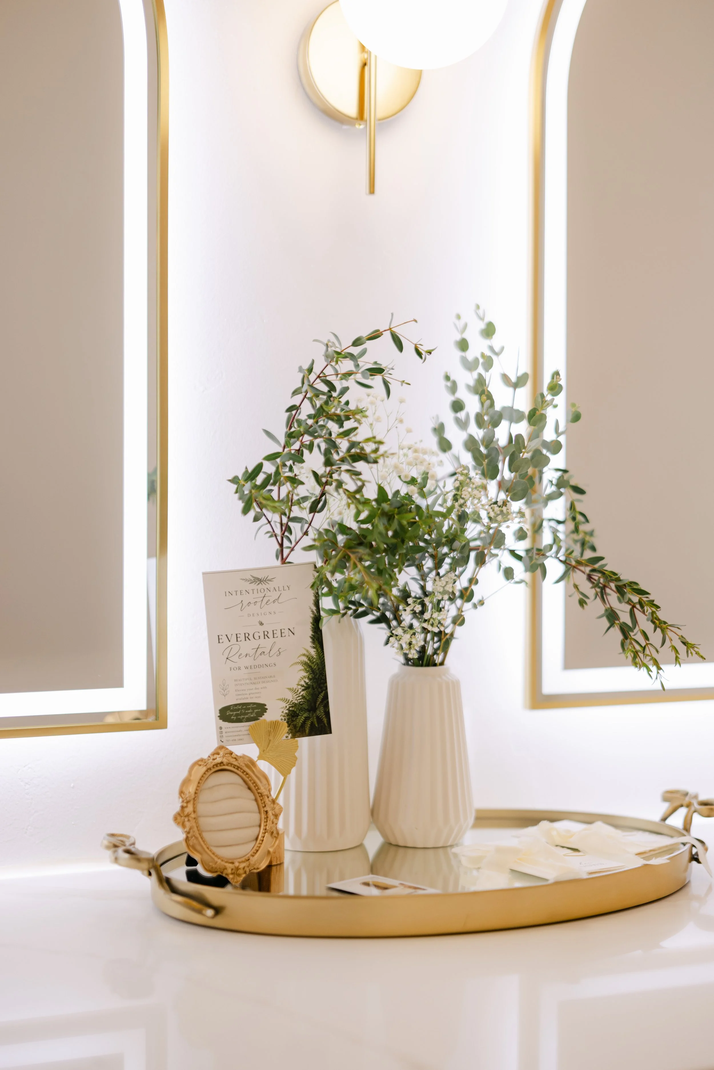 A white table with a gold tray, a decorative gold picture frame, a white vase with greenery and white flowers, and a promotional card for Evergreen Rentals, set against illuminated rectangular and arched mirrors with gold borders on a white wall.