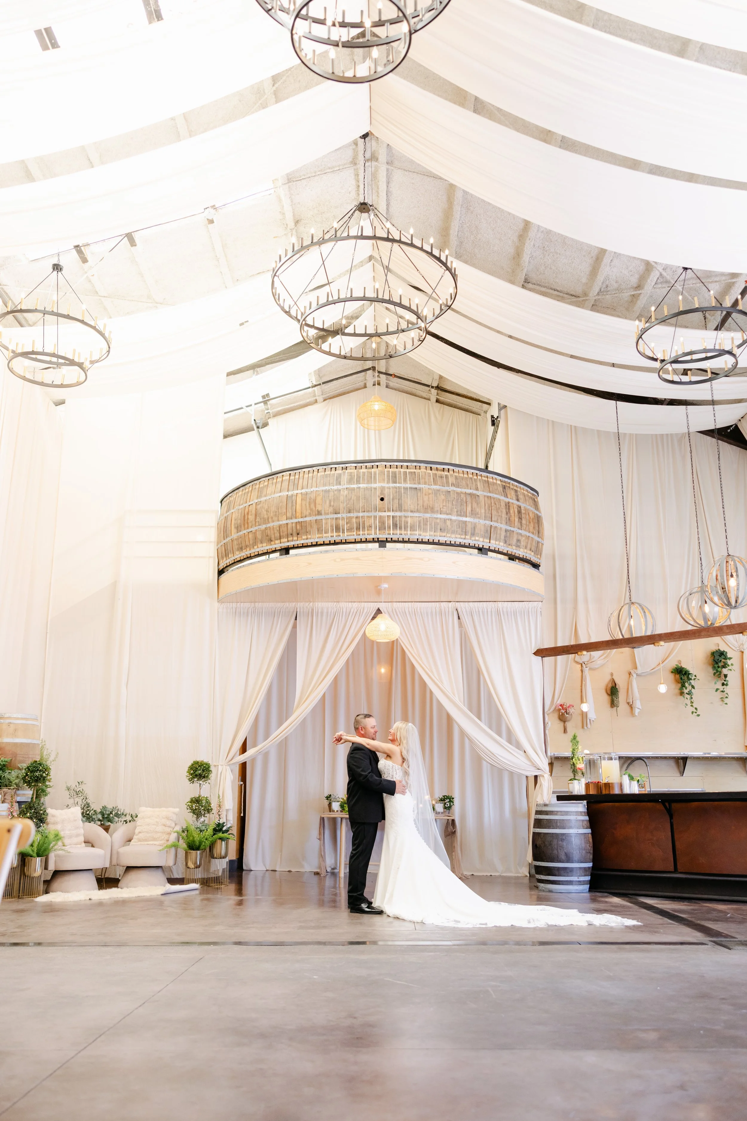 A bride and groom dance in a decorated indoor wedding venue with draped white fabric on the ceiling, chandeliers, and elegant furniture.