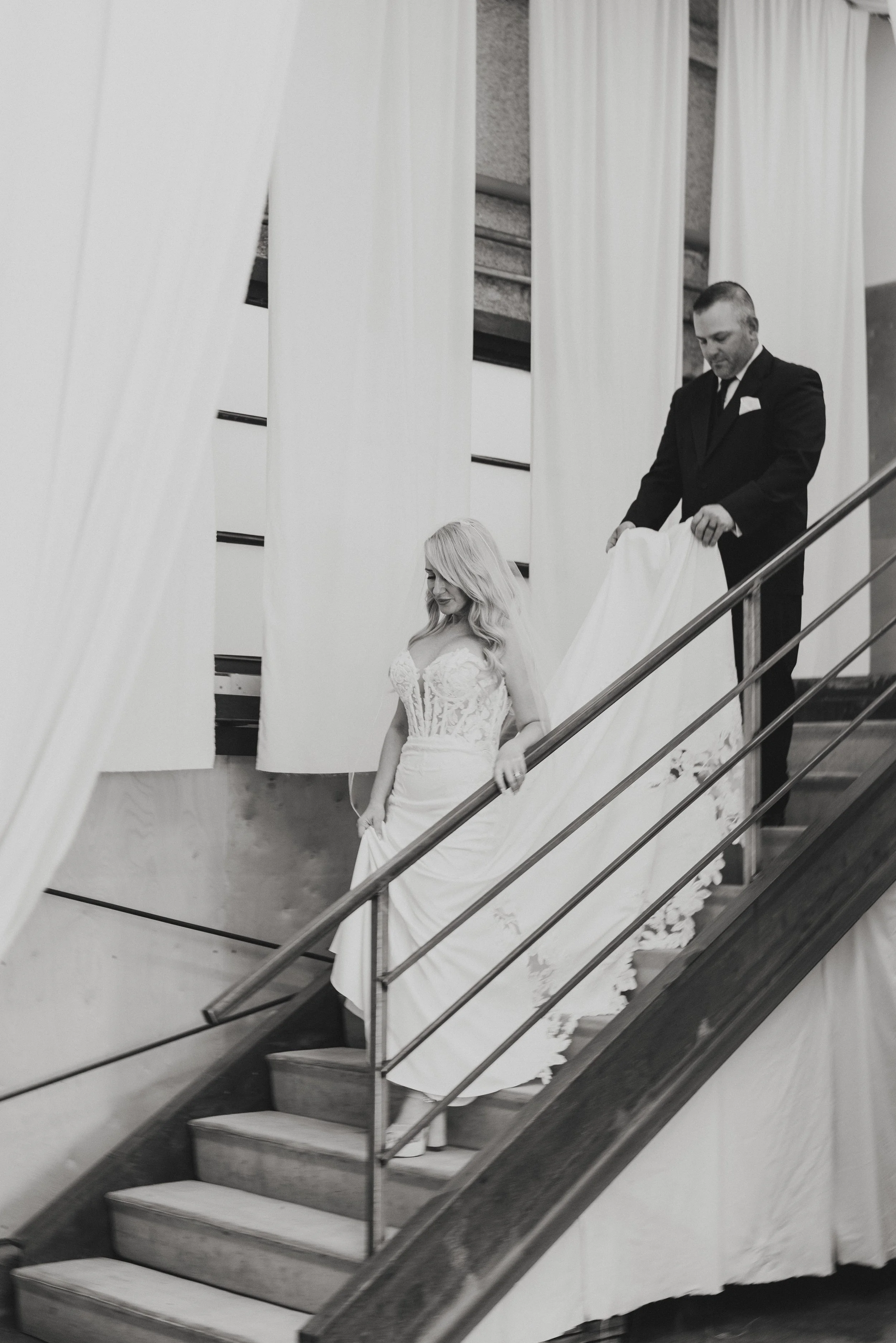 A bride with long blonde hair in a lace wedding gown descending a staircase, with a man in a suit holding her train. The staircase has wooden steps and modern metal railings, and is lined with white curtains.