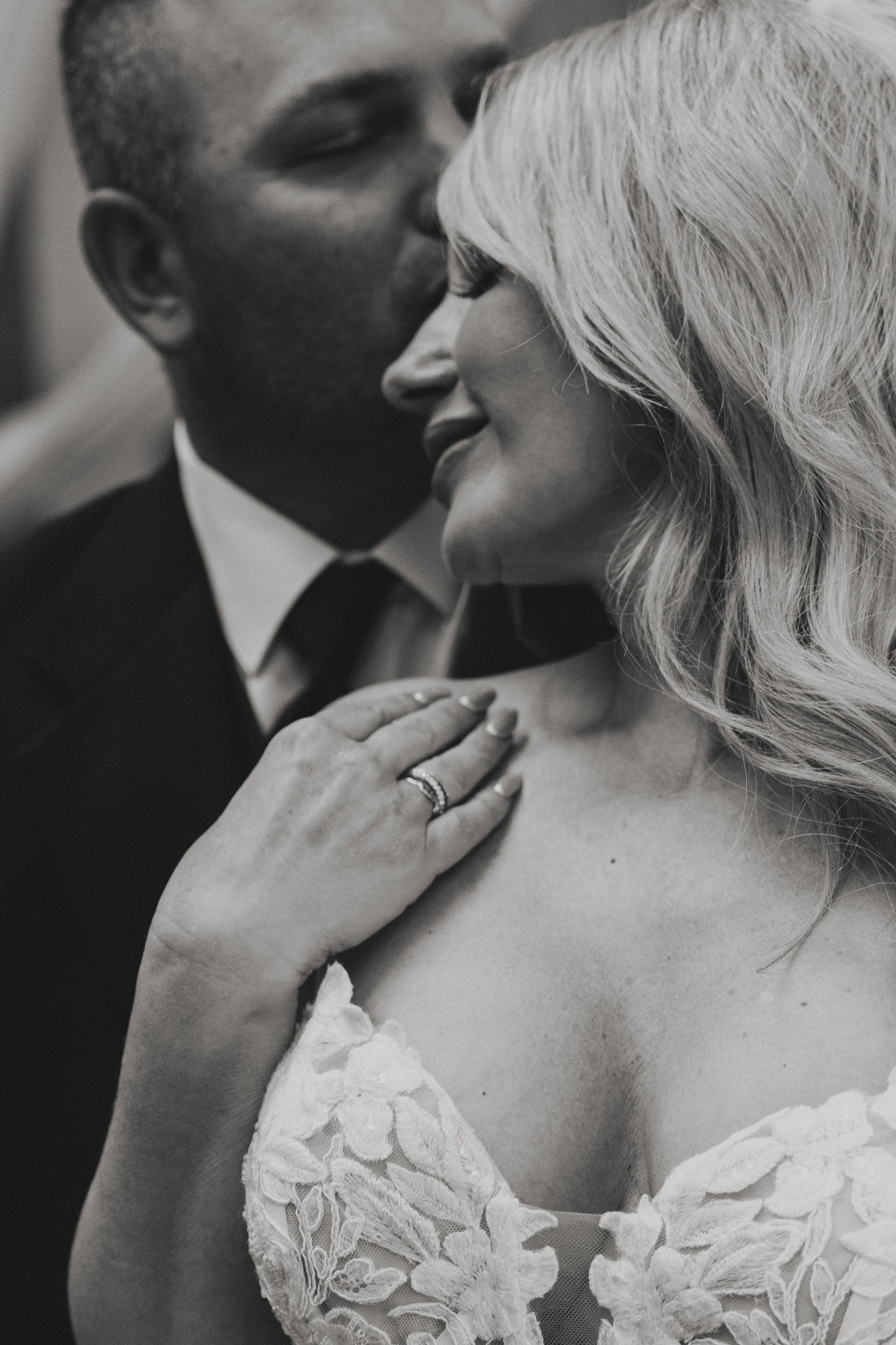 A black and white close-up photo of a couple kissing, focusing on the woman's face and shoulder, with the man's face partially visible. The woman has long, wavy hair and is wearing a white lace garment, possibly a wedding dress. The man's hand is gen