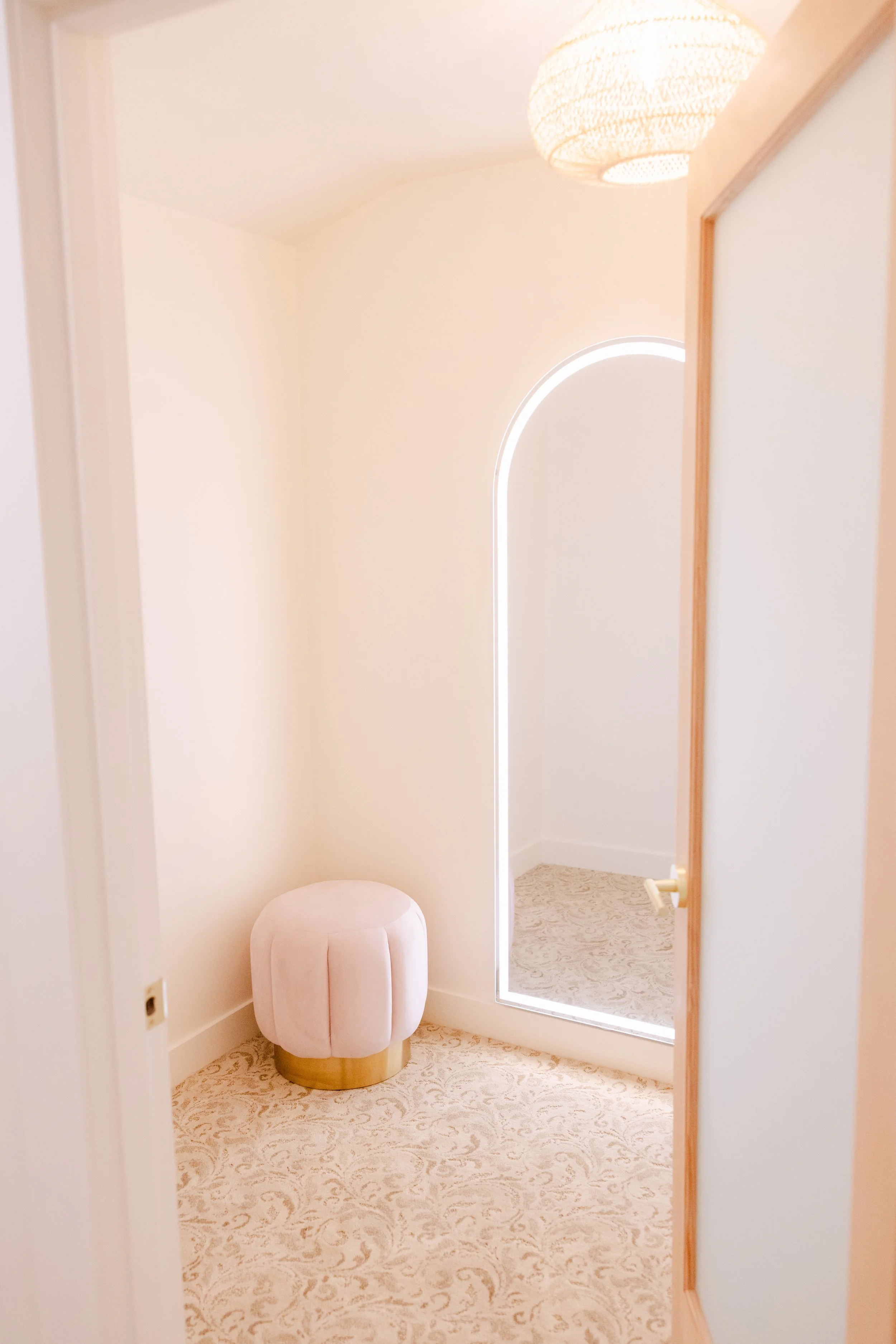 A small, round, blush-colored ottoman with a gold base in a light, cream-colored room. There is a tall mirror with white LED lighting around its edges and a lamp with a woven shade hanging from the ceiling. The room has a patterned carpet.