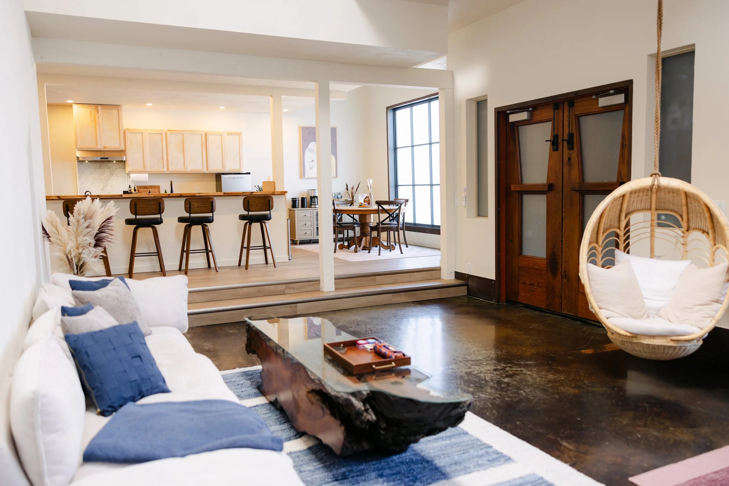 Open-concept living room and kitchen with white walls, wooden accents, and modern furniture. The living room features a white sofa with blue pillows, a wooden coffee table, and a wicker hanging chair with cushions. The kitchen has a breakfast bar wit