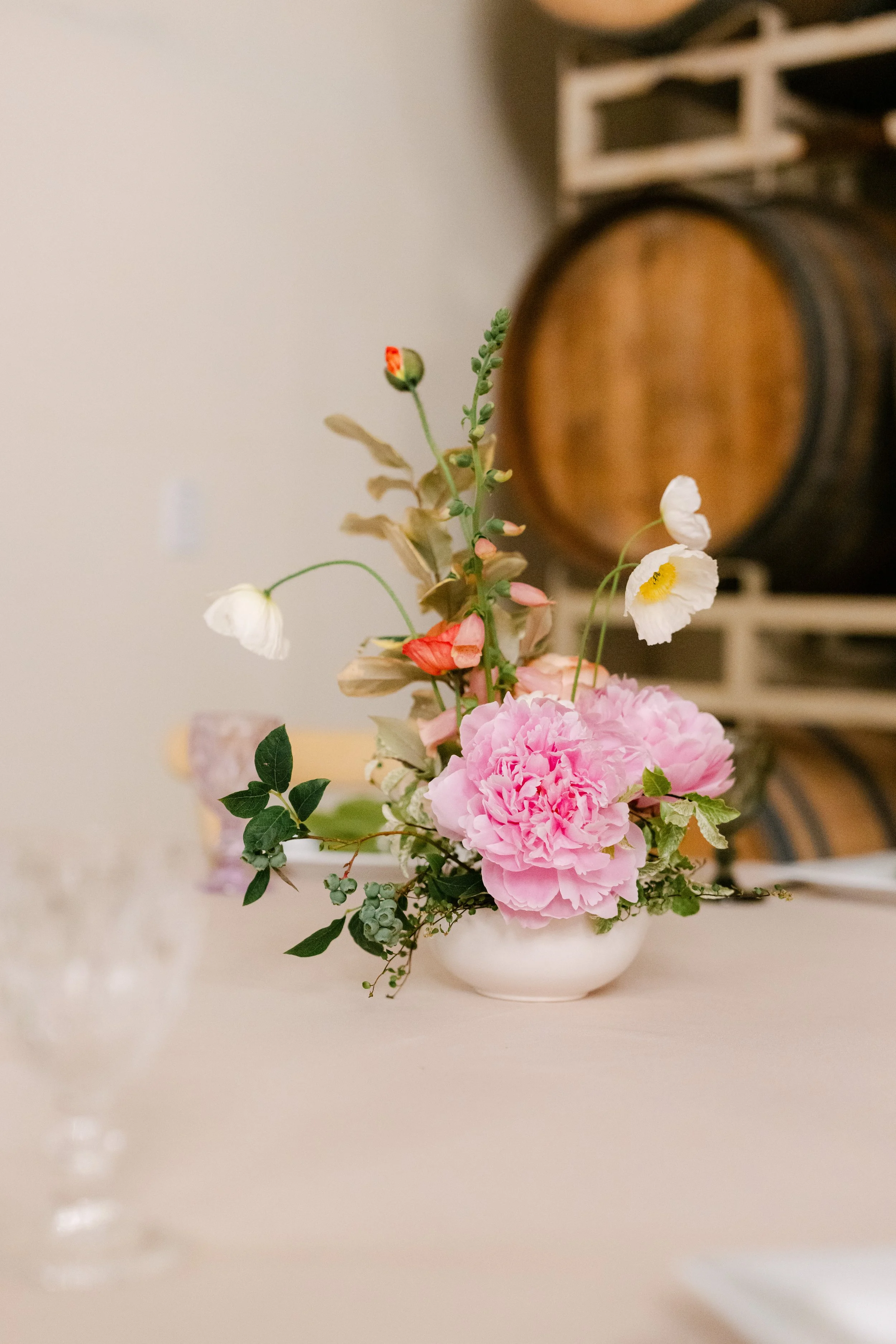 A floral arrangement with pink peonies, white poppies, and green foliage in a white bowl on a light-colored surface, with a blurred background including a wooden barrel and shelving unit.