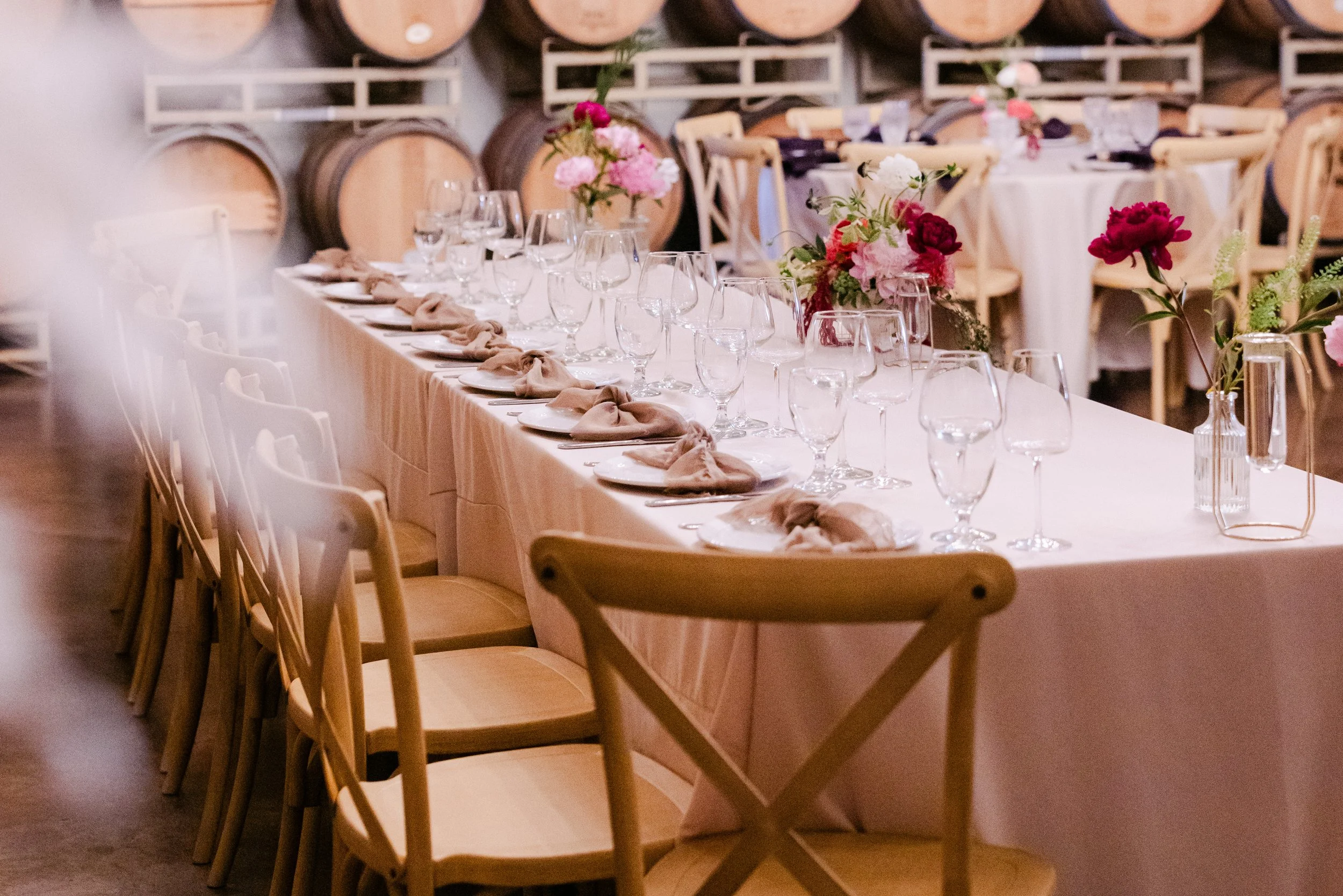 A long banquet table decorated with pink and white flower arrangements, set with plates, napkins, glasses, and cutlery, in a venue with wine barrels and chairs.