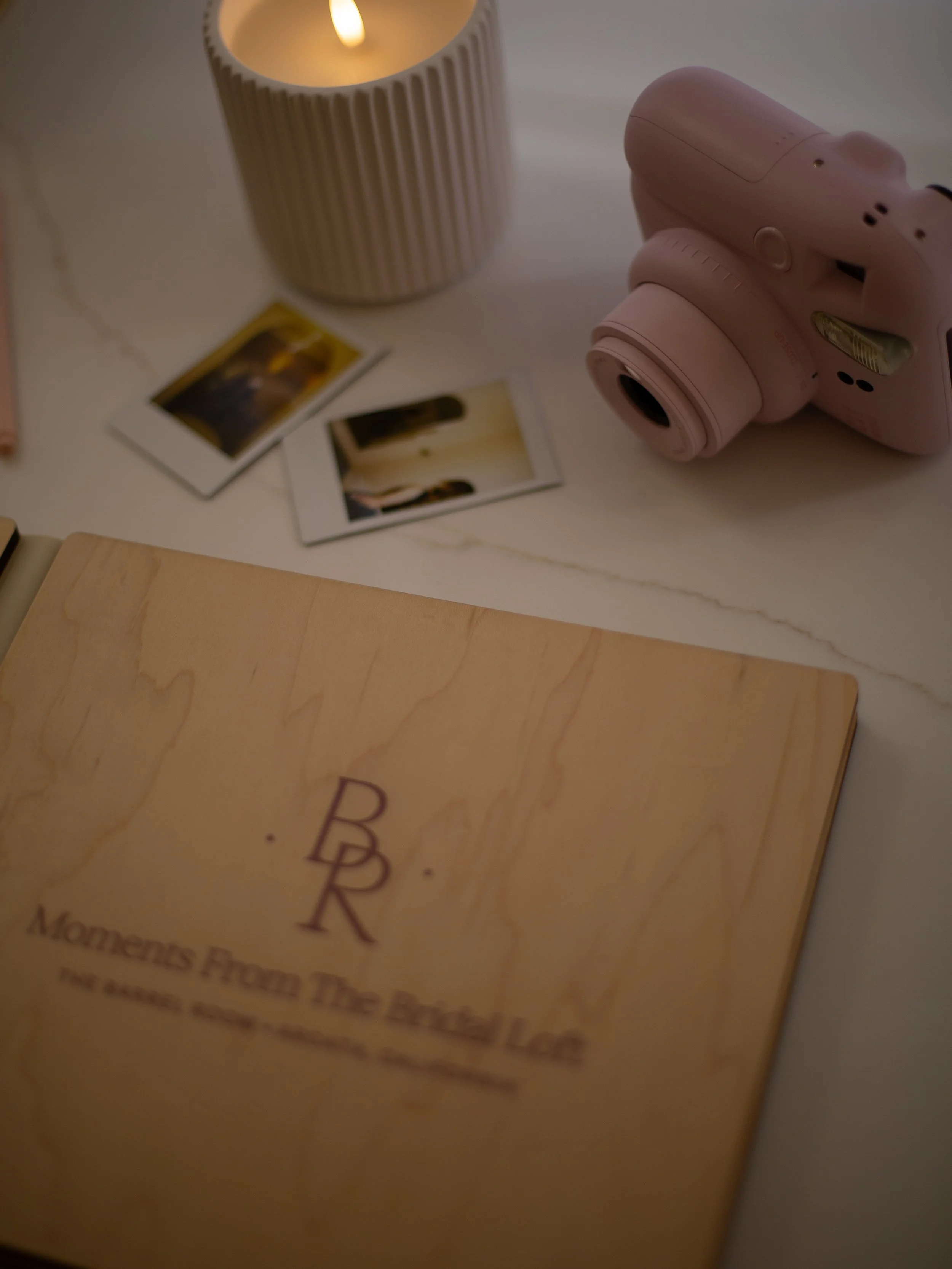 Close-up of a wooden block with embossed letters, a pink camera, a beige ruffled candle holder with a lit candle, and two Polaroid photos on a white surface.