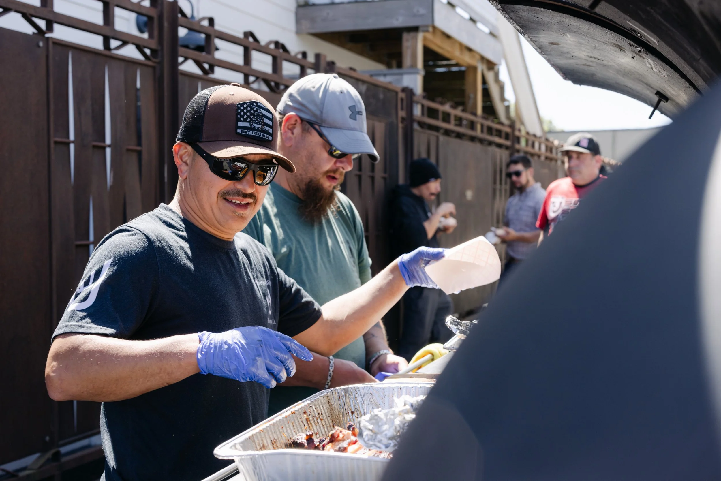 People serving food at an outdoor gathering, with one man wearing sunglasses and a cap, and others in the background, some wearing sunglasses and hats.