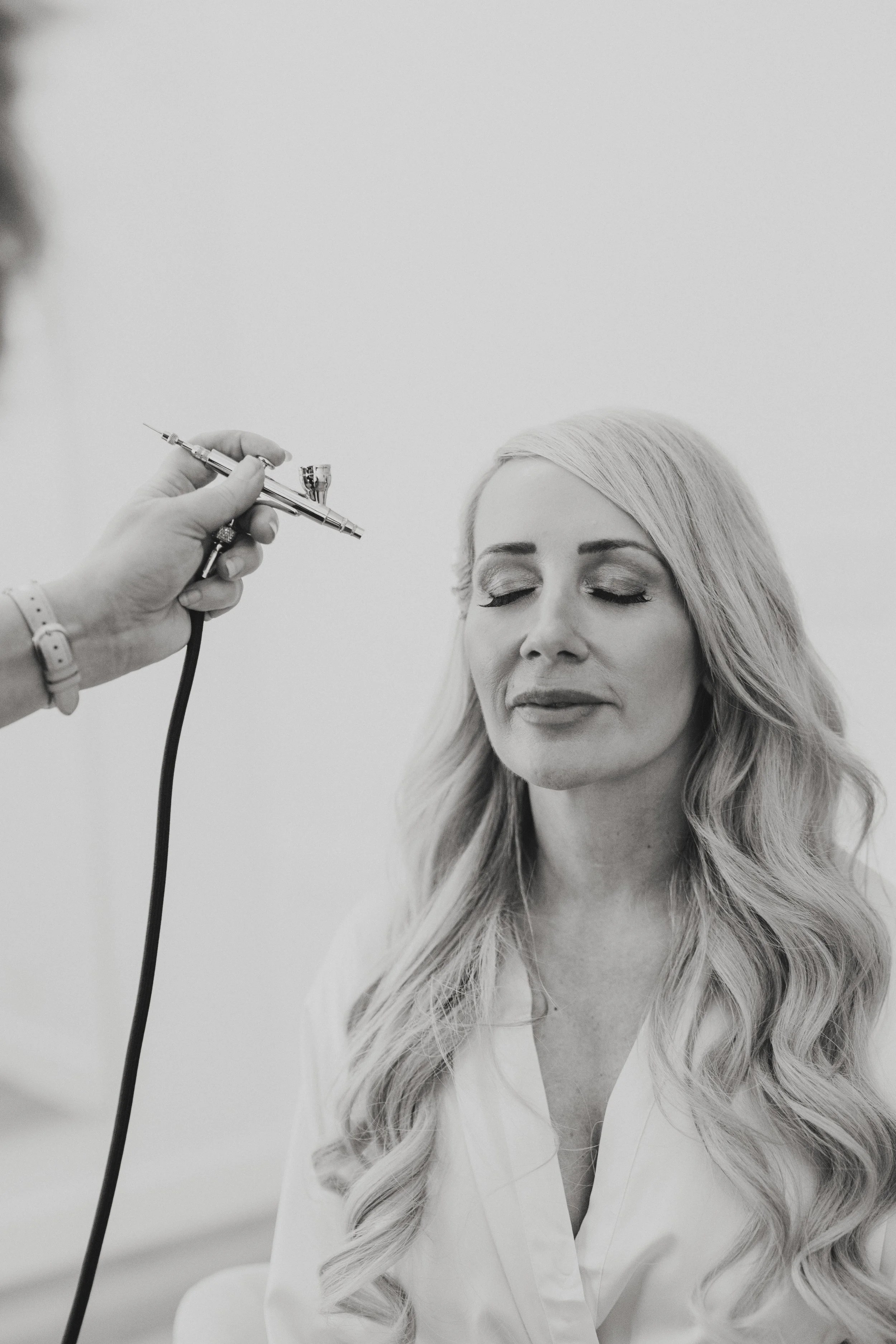 A woman with long, wavy hair having her face treated with a cosmetic syringe by a professional in a clinical setting.