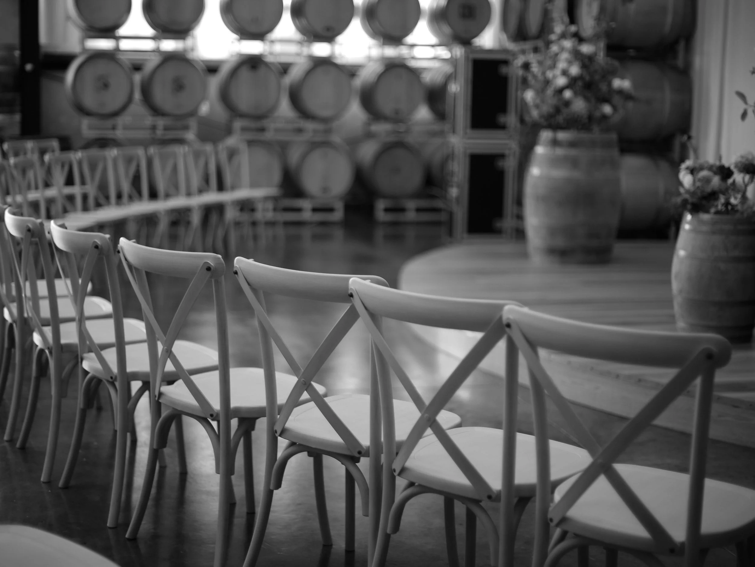 Rows of empty chairs facing a wall of wine barrels in a winery, with some flowers in large vases to the side.