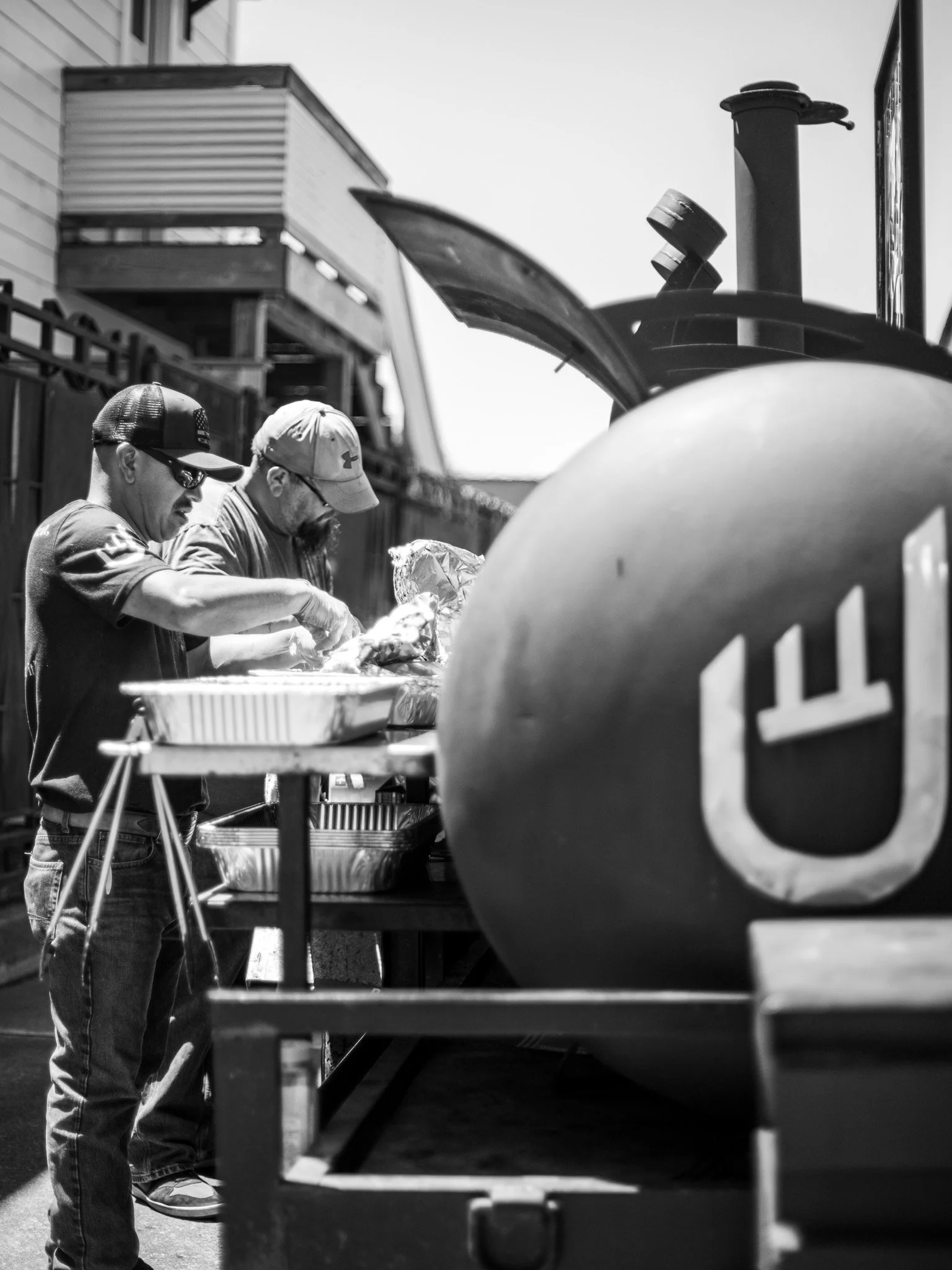 Two men working at an outdoor grill station, with a large grill in front and a building with a balcony in the background. One man wears a cap and sunglasses, the other a cap and glasses.