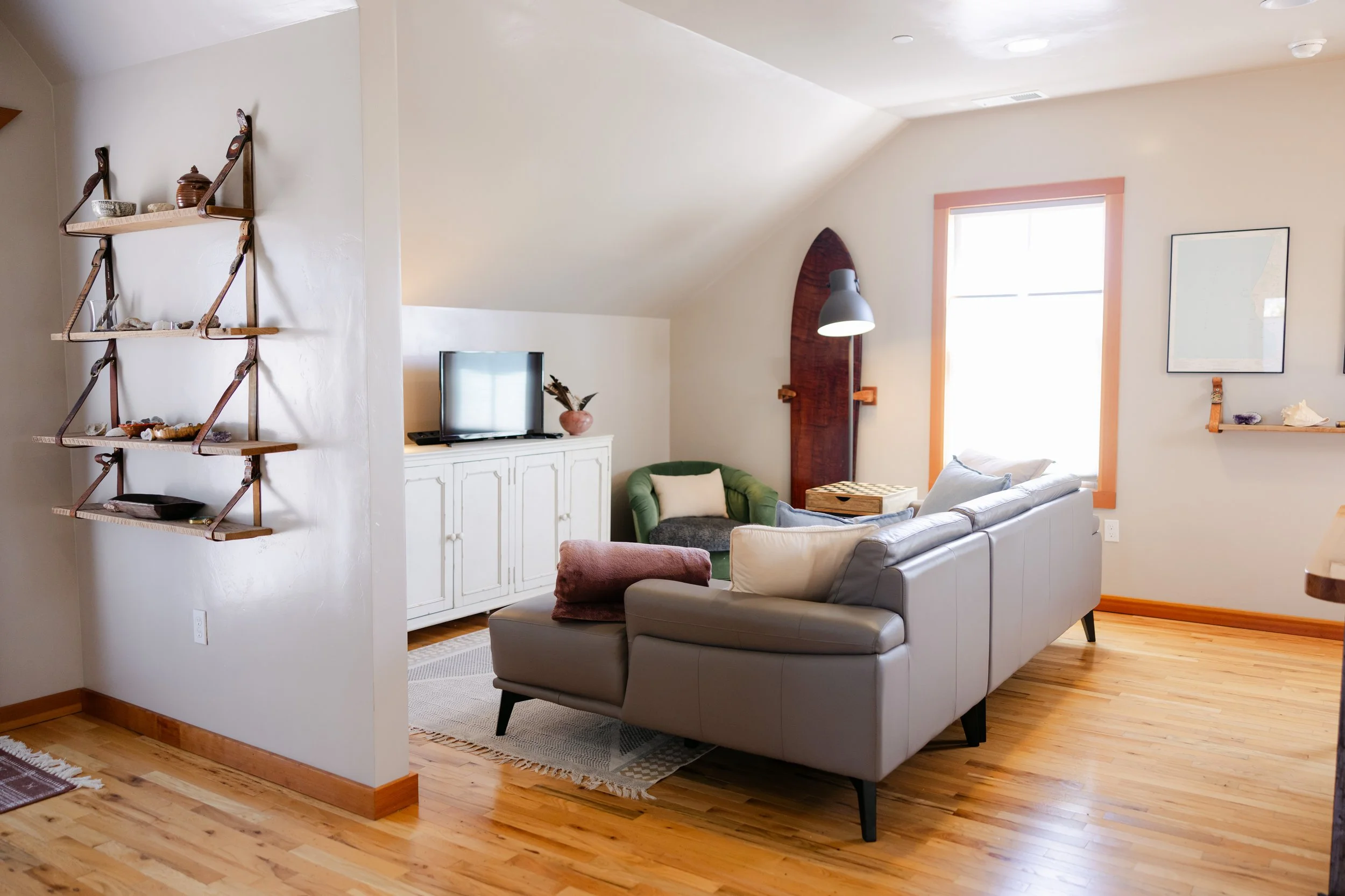 A cozy living room with a white couch, a green armchair, a white cabinet with a TV on top, a window with pink trim, and wooden floors.
