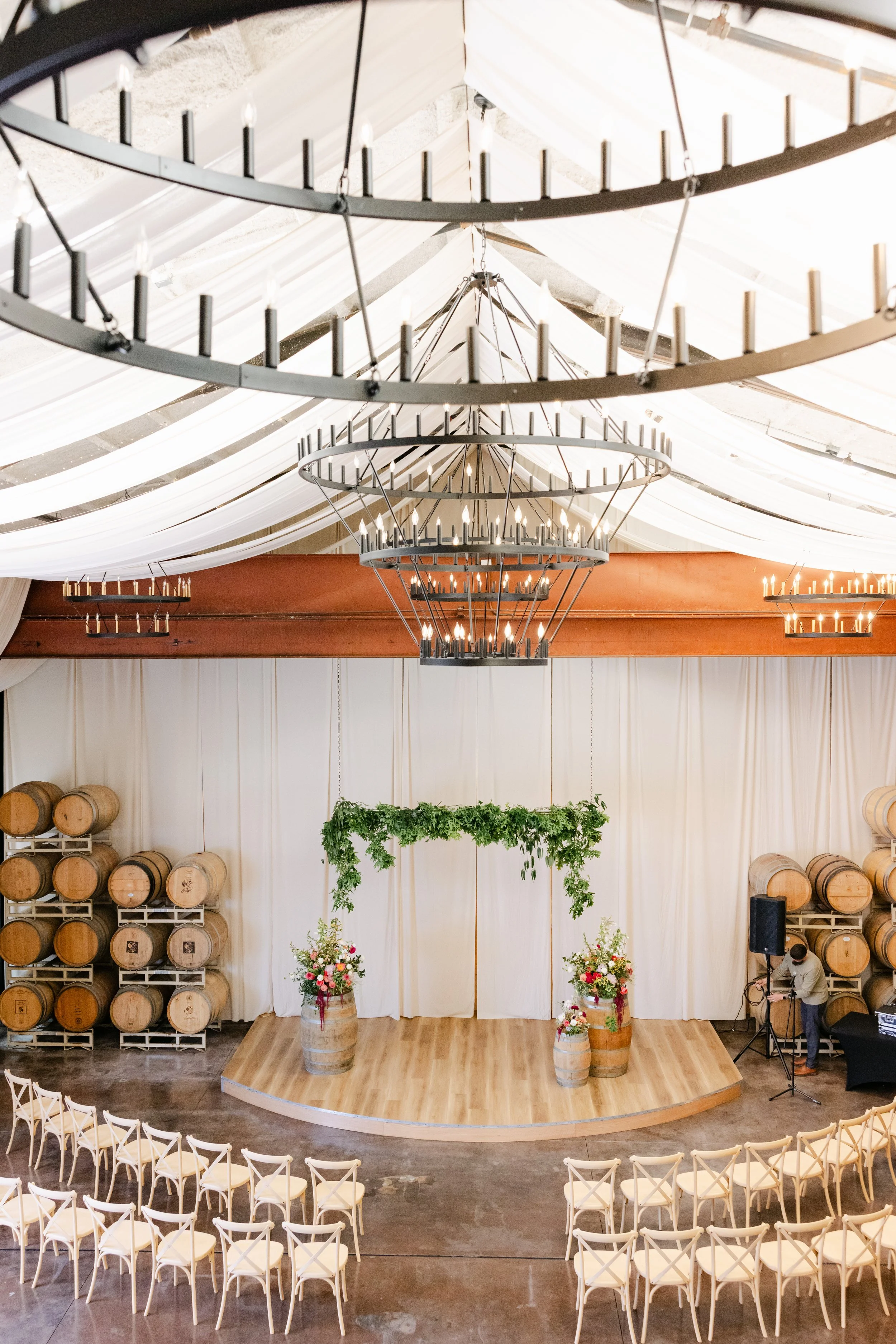 Wedding ceremony setup in a large hall with rows of white chairs arranged facing a small stage with floral arrangements and hanging greenery. There are several large circular chandeliers hanging from the ceiling, which is decorated with white drapery